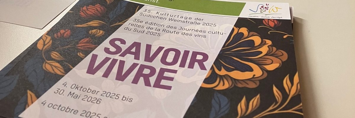 Savoir vivre, so lautete das Motto der SÜW-Kulturtage 2025. Foto: KV SÜW Flyer mit entsprechender Aufschrift liegt auf einem Stehtisch, im Hintergrund großformatige Kunstwerke an einer weißen Wand.