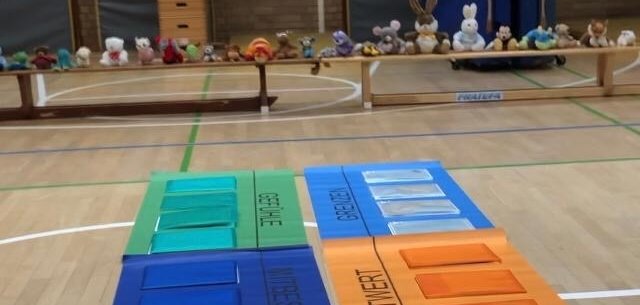 Auf dem Boden liegen sechs bunte Plakate mit verschiedenen Aufgaben-Taschen darauf. Im Hintergrund steht eine lange Bank mit Kuscheltieren darauf. 