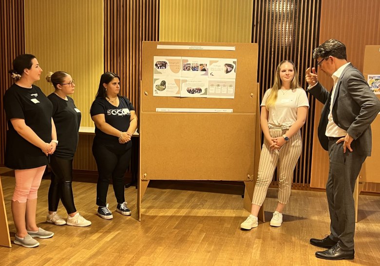 Staatssekretär Michael Hauer (rechts) im Gespräch mit den SÜW-Auszubildenden in Mainz. Foto: Energieagentur Rheinland-Pfalz Vier junge Frauen und ein Mann vor einer Stellwand.