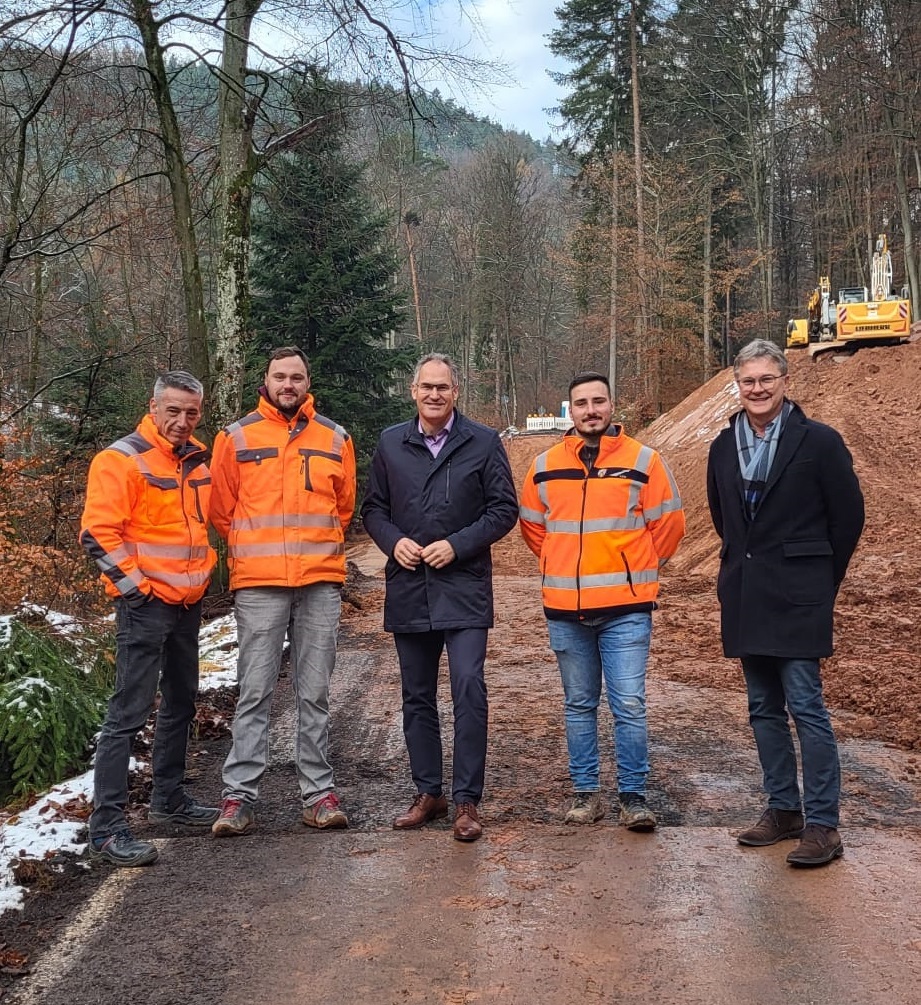 Landrat Dietmar Seefeldt, Mitarbeiter der Firma Gerst und Juchem sowie der Straßenmeisterei Landau und Abteilungsleiter Günter Jung stehen vor der Baustelle der Kreisstraße 6, zu sehen sind eine aufgebrochene Straße sowie ein Erdhügel. 