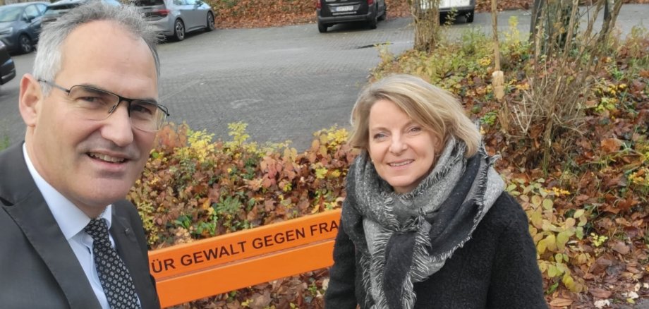 Landrat Dietmar Seefeldt und SÜW-Gleichstellungsbeauftragte Isabelle Stähle stehen vor einer orange gestrichenen Sitzbank am Kreishaus mit Aufschrift „Kein Platz für Gewalt gegen Frauen“. 