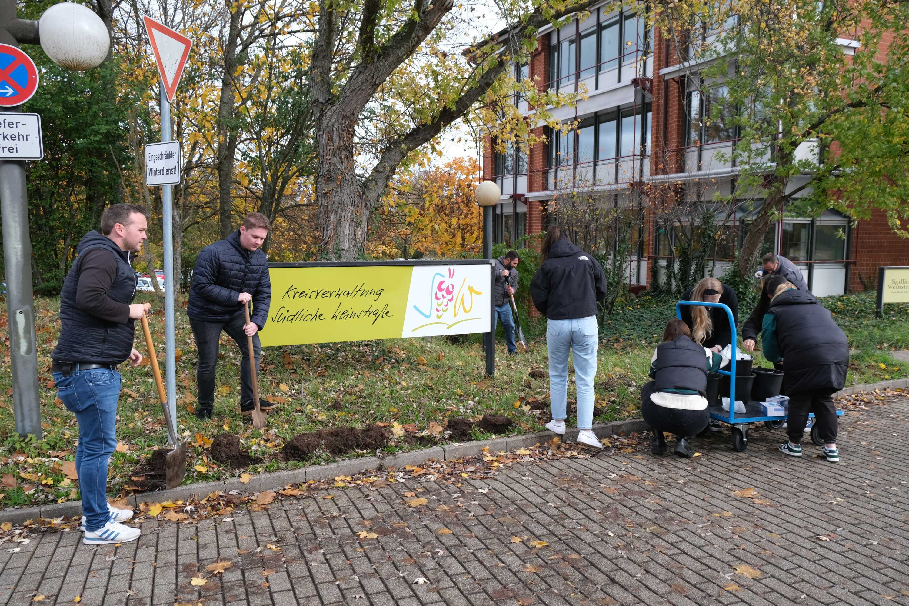 Auszubildende und Verwaltungsmitarbeiter graben Löcher in den Grünstreifen rings um das Willkommen-Schild der Kreisverwaltung.