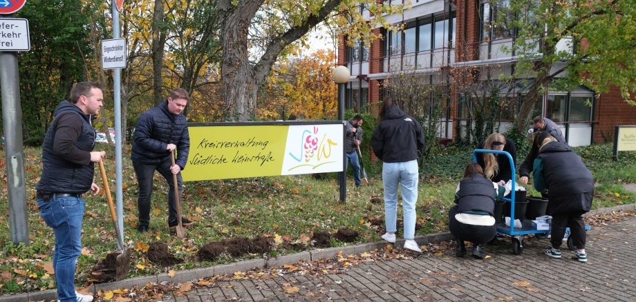 Auszubildende und Verwaltungsmitarbeiter graben Löcher in den Grünstreifen rings um das Willkommen-Schild der Kreisverwaltung.