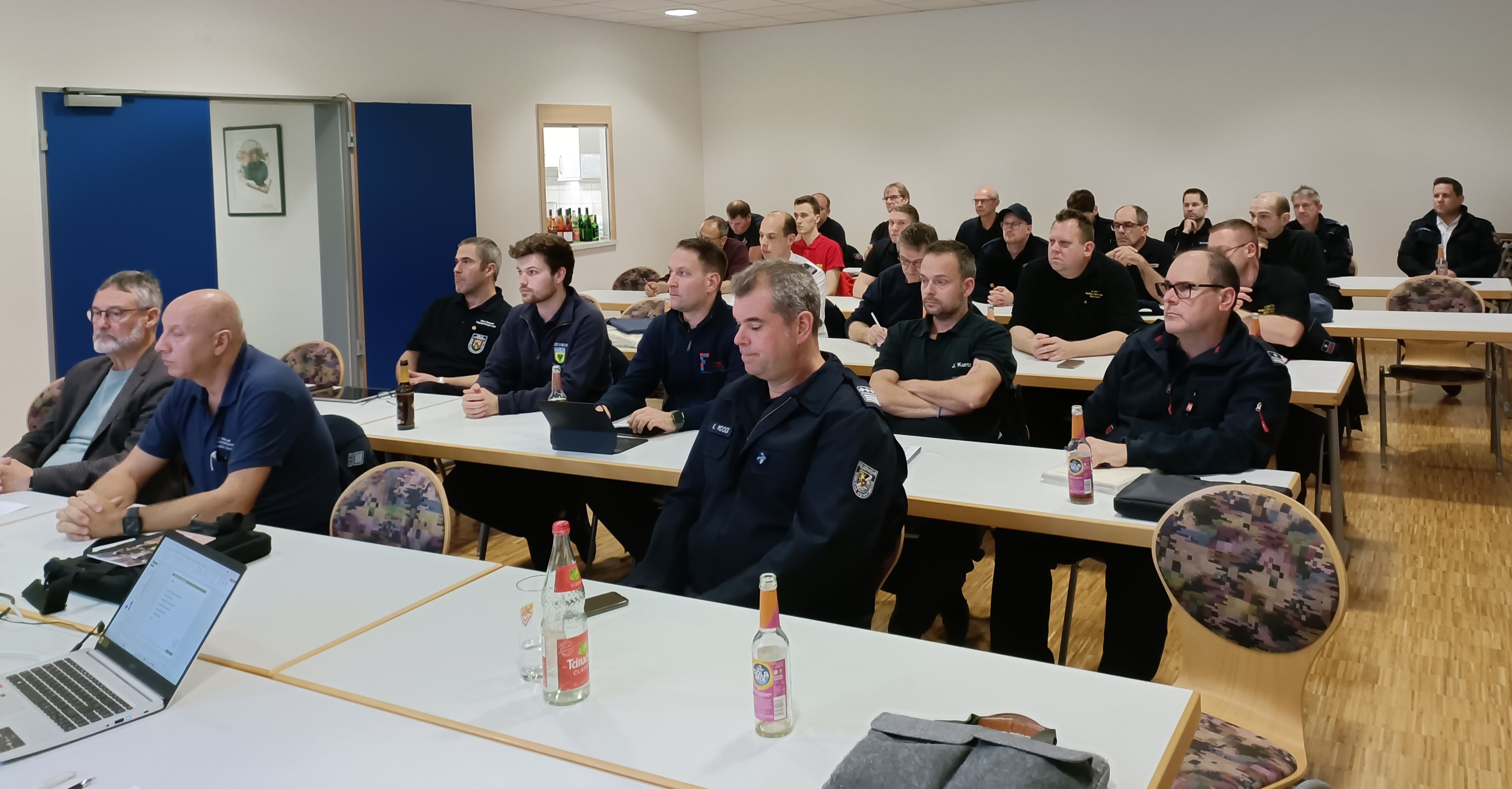 In einem Raum sitzen zahlreiche Männer in Uniform an aufgestellten Tischreihen. 