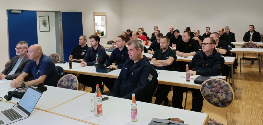 In einem Raum sitzen zahlreiche Männer in Uniform an aufgestellten Tischreihen. 