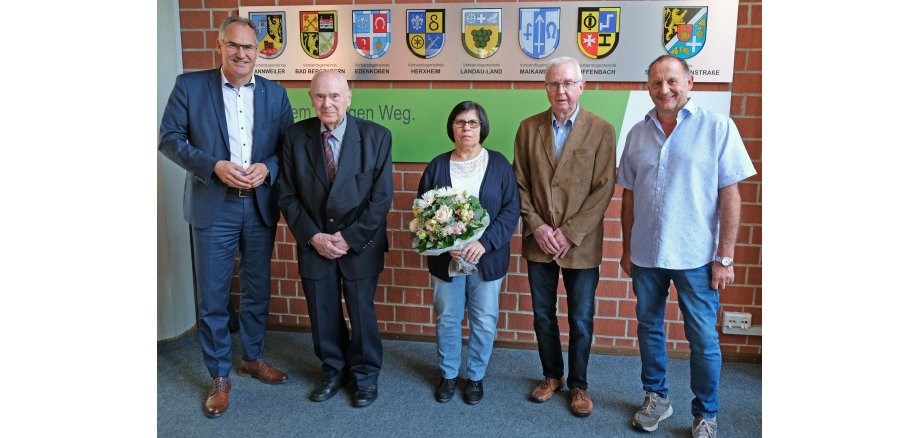 Fünf Personen stehen vor einer Wand mit Wappen. 