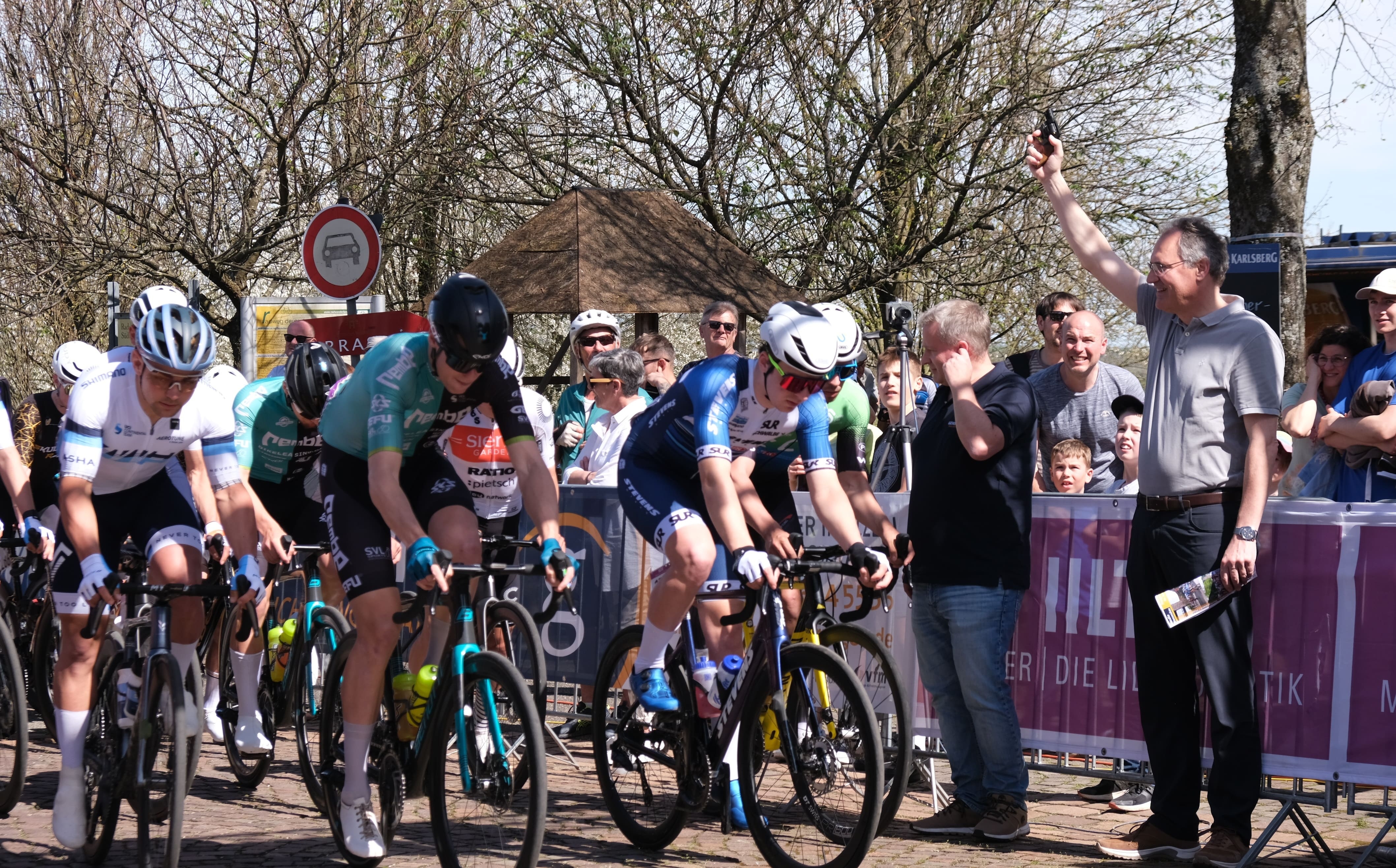 Landrat Dietmar Seefeldt, rechts im Bild, gibt den Startschuss für den Großen Preis der Südlichen Wienstraße. Links sind einige Radfahrer zu sehen, die das Rennen Starten. Im Hintergrund stehen hinter Absperrungen zahlreiche Zuschauer.