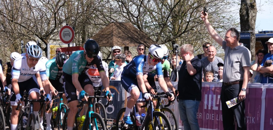 Landrat Dietmar Seefeldt, rechts im Bild, gibt den Startschuss für den Großen Preis der Südlichen Wienstraße. Links sind einige Radfahrer zu sehen, die das Rennen Starten. Im Hintergrund stehen hinter Absperrungen zahlreiche Zuschauer.
