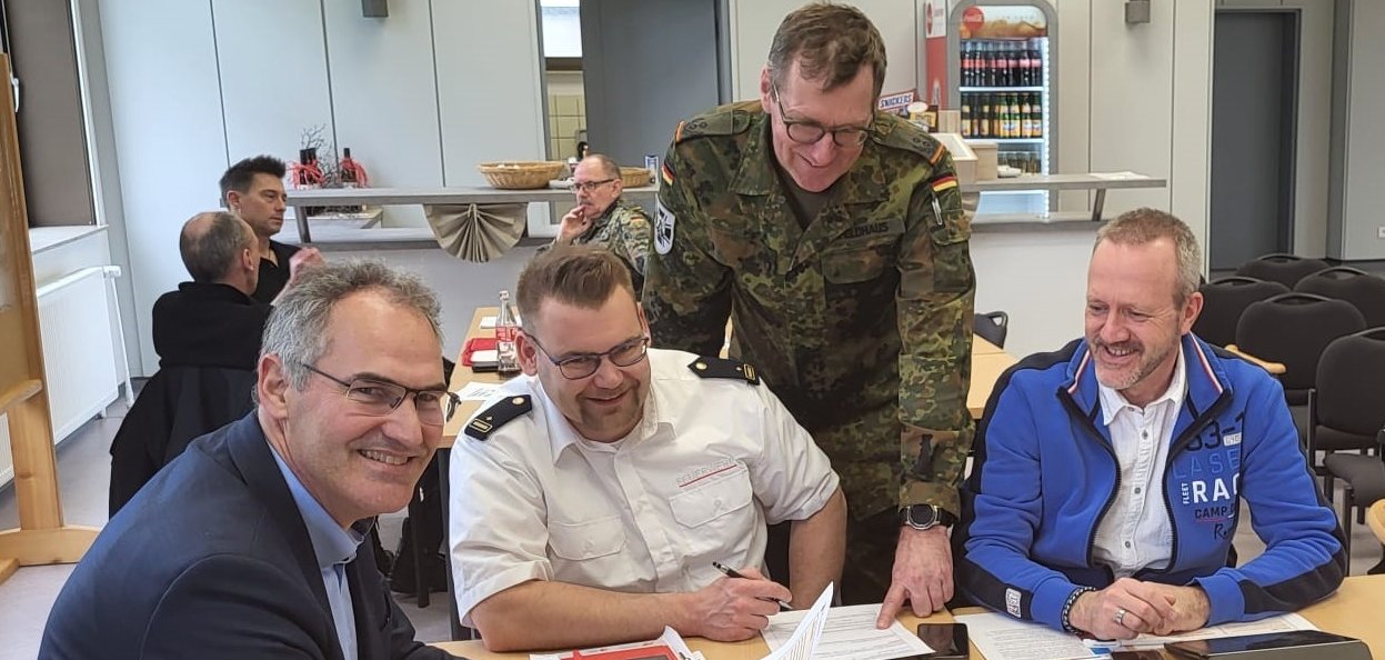 Landrat Dietmar Seefeldt hat sich bei der zweitägigen Stabsrahmenübung in Mainz im Gespräch mit SÜW-Brand- und Katastrophenschutzinspekteur Jens Thiele, Oberstleutnant Joachim Feldhaus vom Kreisverbindungskommando SÜW und dem Leiter des SÜW-Verwaltungsstabs Peter Schürmann einen Überblick über das Übungsgeschehen verschafft. Sie alle sitzen gemeinsam an einem Tisch mit Dokumenten darauf. Foto: KV SÜW