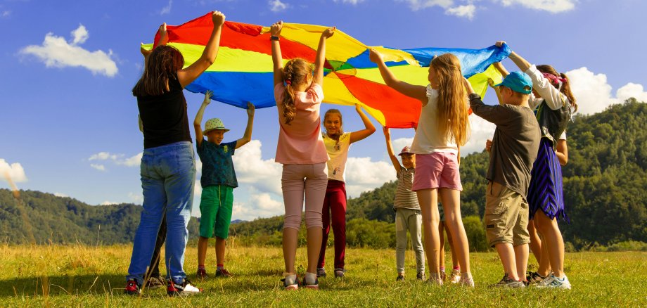 Kinder und Jugendliche stehen bei schönem Wetter im Kreis auf einer Wiese und halten dabei ein buntes Tuch, dass sie für ein Spiel verwenden.