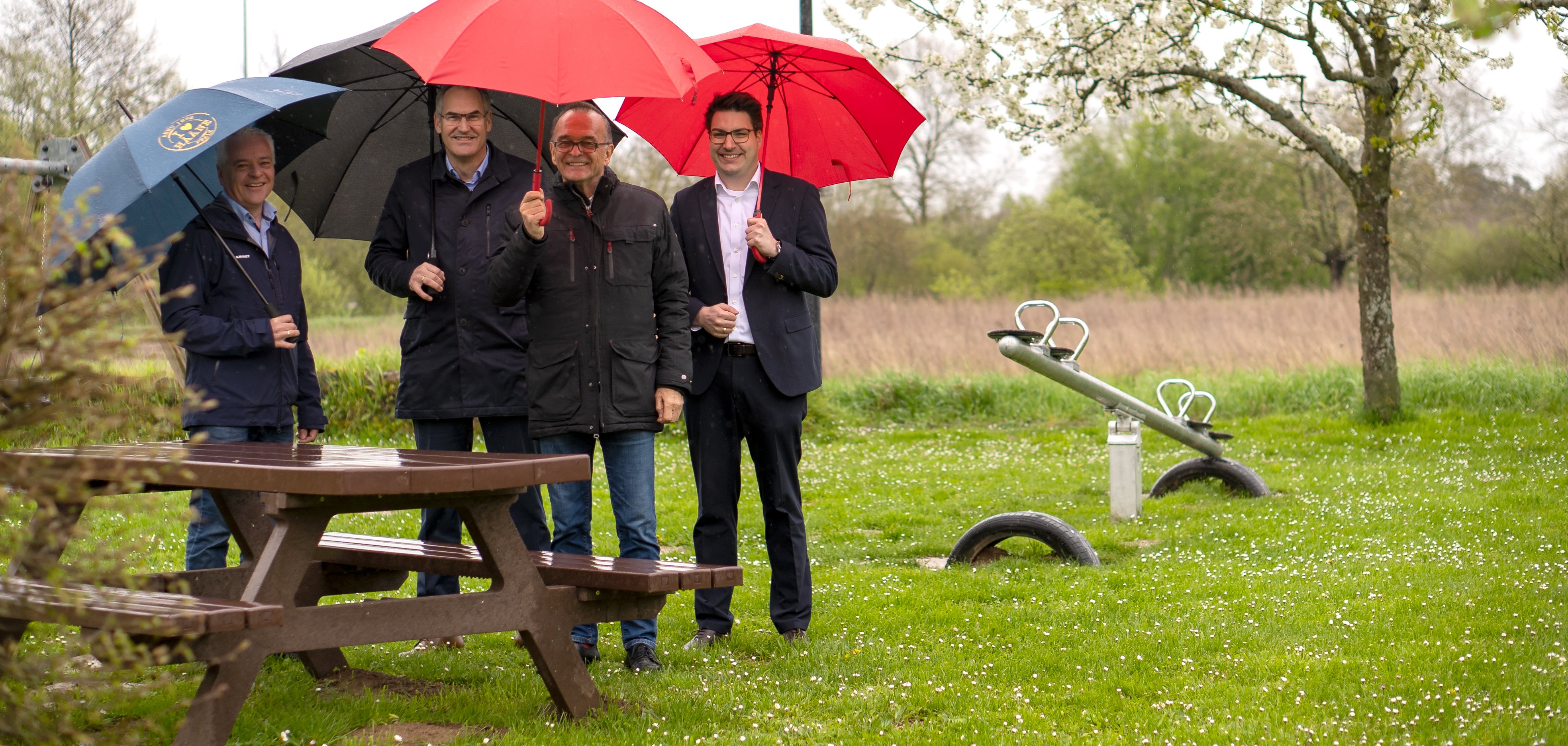 Haynas Ortsvorsteher Markus Dudenhöffer, Landrat Dietmar Seefeldt (Kreis SÜW), Landrat Dr. Fritz Brechtel (Kreis GER) und Benjamin Hirsch, Vorstandsmitglied der Sparkasse Südpfalz, stehen im Regen mit aufgespannten Schirmen neben der Sitzgruppe auf dem Spielplatz in Hanya.