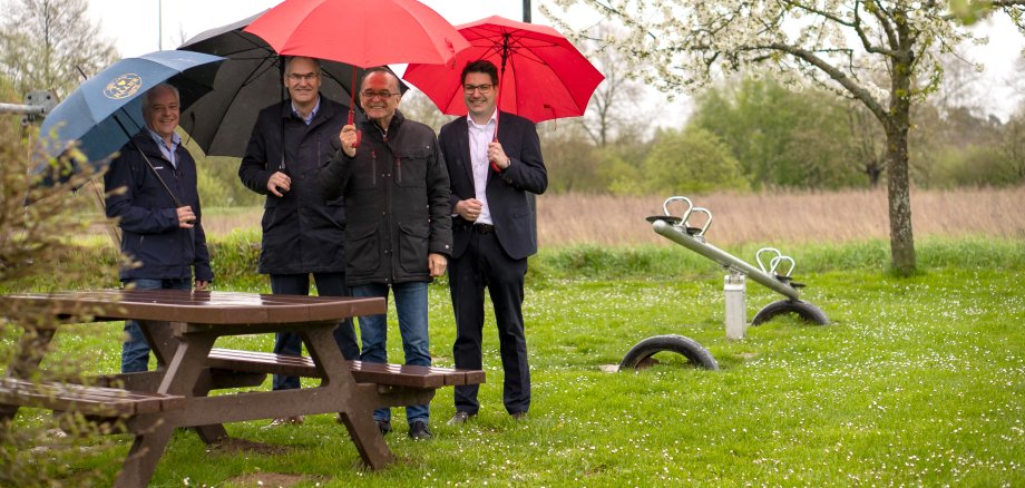 Haynas Ortsvorsteher Markus Dudenhöffer, Landrat Dietmar Seefeldt (Kreis SÜW), Landrat Dr. Fritz Brechtel (Kreis GER) und Benjamin Hirsch, Vorstandsmitglied der Sparkasse Südpfalz, stehen im Regen mit aufgespannten Schirmen neben der Sitzgruppe auf dem Spielplatz in Hanya.