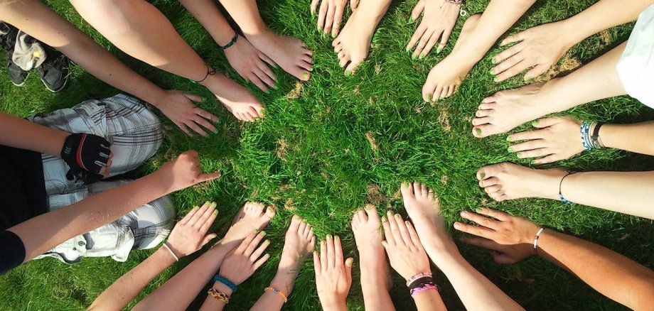 Eine Gruppe Jugendlicher platziert Hände und Füße im Kreis auf einer Wiese.