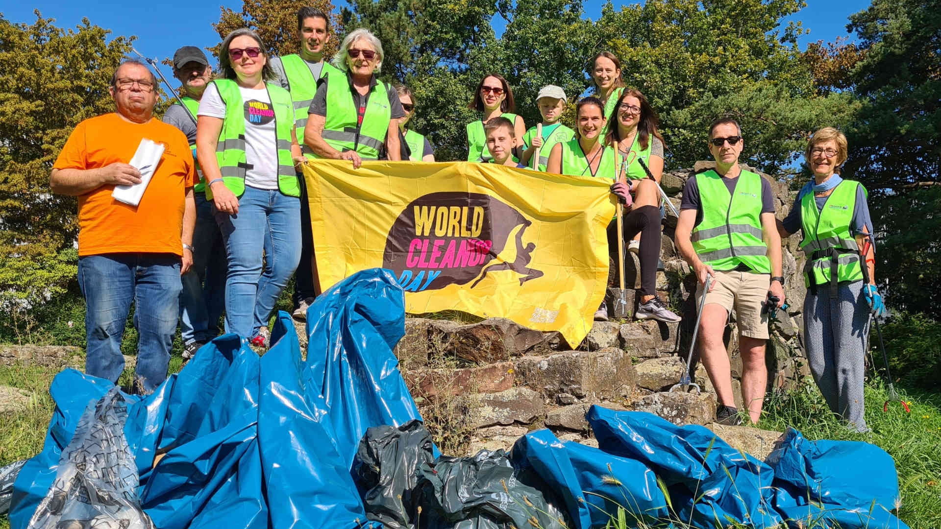 Teilnehmerinnen und Teilnehmer des World Clean Up Days 2023 halten ein gelbes Banner mit der Aufschrift "World Clean Up Day". Die Personen tragen Warnwesten und halten teils Greifzangen zum Sammeln von Müll in der Hand. Vor ihnen liegen einige blaue Müllsäcke.