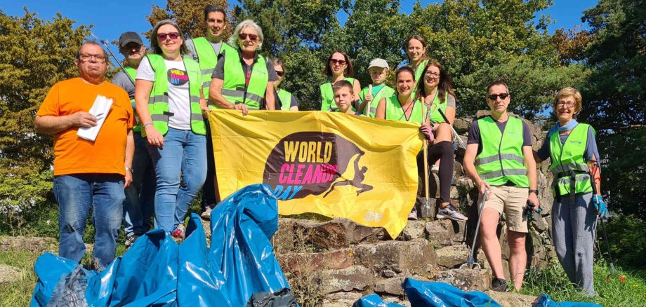 Teilnehmerinnen und Teilnehmer des World Clean Up Days 2023 halten ein gelbes Banner mit der Aufschrift "World Clean Up Day". Die Personen tragen Warnwesten und halten teils Greifzangen zum Sammeln von Müll in der Hand. Vor ihnen liegen einige blaue Müllsäcke.