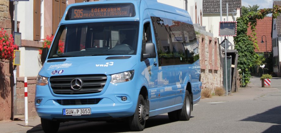 Ein blauer Kleinbus steh an einer Haltestelle innerhalb einer Ortschaft. Im Hintergrund sind Wohnhäuser und eine Kirche zu sehen.