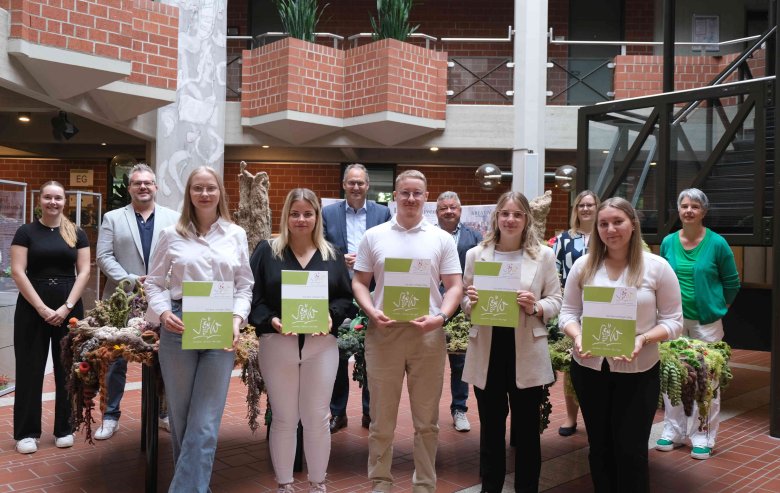 Hanna Klein, Michelle Fösser, Leon Frankfurter, Sophie Klatz und Laura Müller (erste Reihe von links) haben das duale Studium „Verwaltung“ begonnen. In der hinteren Reihe stehen Personalsachbearbeiterin Anna Noltze, Sebastian Renner, Referatsleiter „Personal und Organisationsentwicklung, Zentrale Dienste“, Landrat Dietmar Seefeldt, Personalratsvorsitzender Udo Baumgartner, Larissa Conrath, Sachgebietsleiterin Personal, und Gisela Nist, interne Gleichstellungsbeauftragte (von links). Foto: KV SÜW Eine Gruppe von Menschen steht im Eingangsbereich der Kreisverwaltung Südliche Weinstraße.