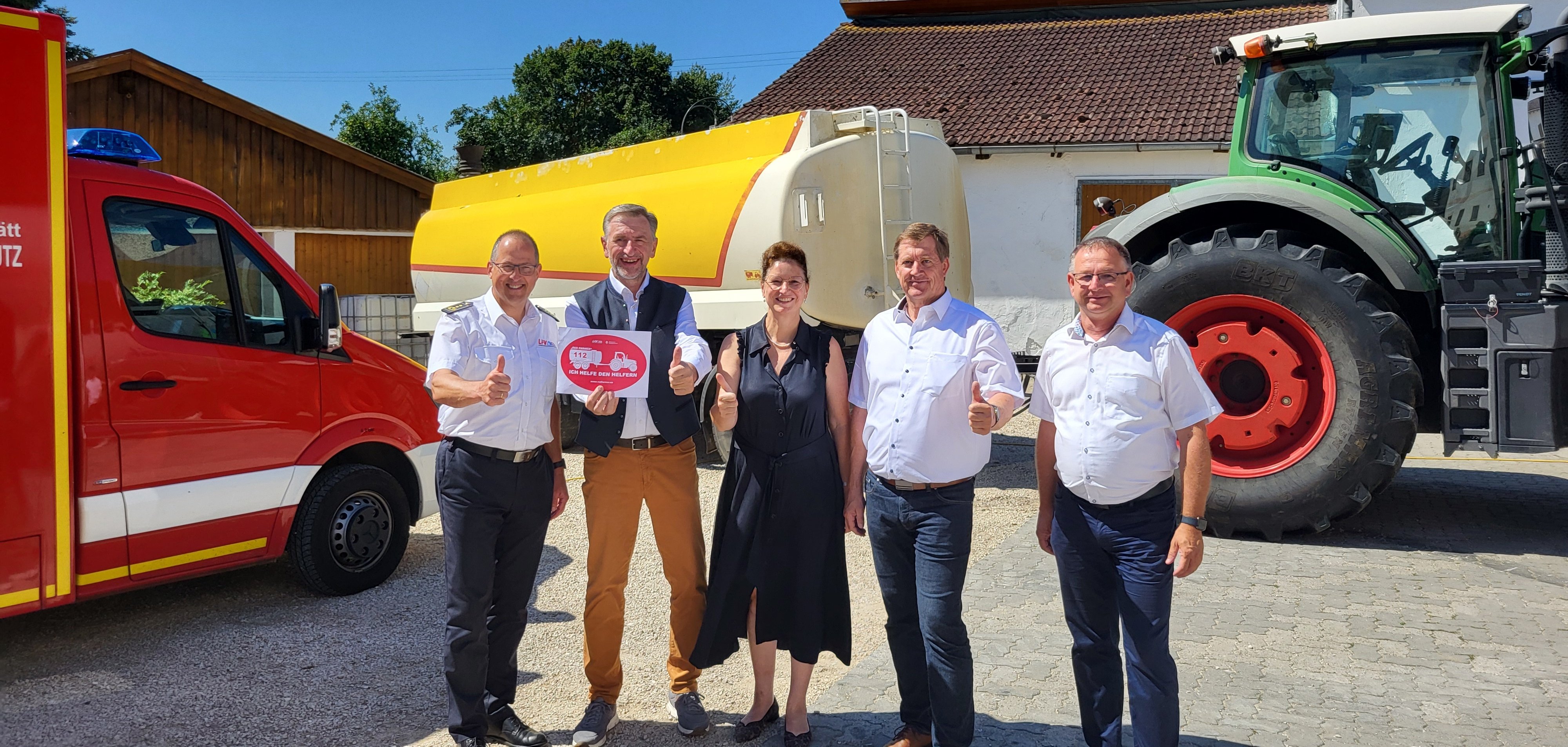 Fünf Vertreterinnen und Vertreter des Bayerischen Bauernverbands und des Landesfeuerwehrverbands Bayern stehen gemeinsam vor einem Feuerwehrfahrzeug, einem Wassertankwagen und einem Traktor. Sie lächeln und zeigen den Daumen nach oben, während sie den Start des Kooperationsprojekts 'Red Farmer' feiern. Unter ihnen ist der Herxheimer Landwirt Thomas Knecht (ganz rechts). Im Hintergrund sind landwirtschaftliche Gebäude und ein klarer, blauer Himmel zu sehen.