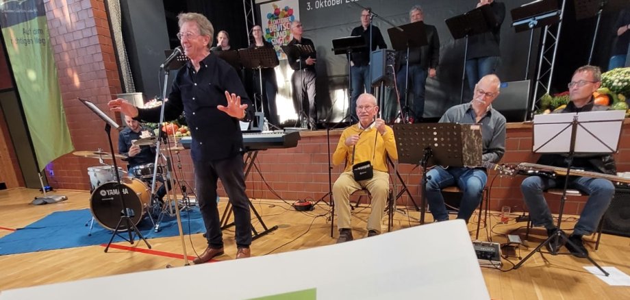 Auf dem Bild ist eine musikalische Veranstaltung im Rahmen der Aktion „Deutschland singt und klingt“ zu sehen, die am 3. Oktober 2022 in der Wiesentalhalle Steinfeld stattfand. Im Vordergrund wird ein Liederheft mit dem Titel „Pfälzer Wind“ gezeigt, während im Hintergrund eine Live-Band unter der Leitung eines Dirigenten auf der Bühne musiziert. Einige Chormitglieder singen dazu. Die Veranstaltung diente dem gemeinsamen Singen zur Feier des Tages der Deutschen Einheit. Ein Banner im Hintergrund trägt den Text „Auf dem richtigen Weg. Auch im geeinten Deutschland.