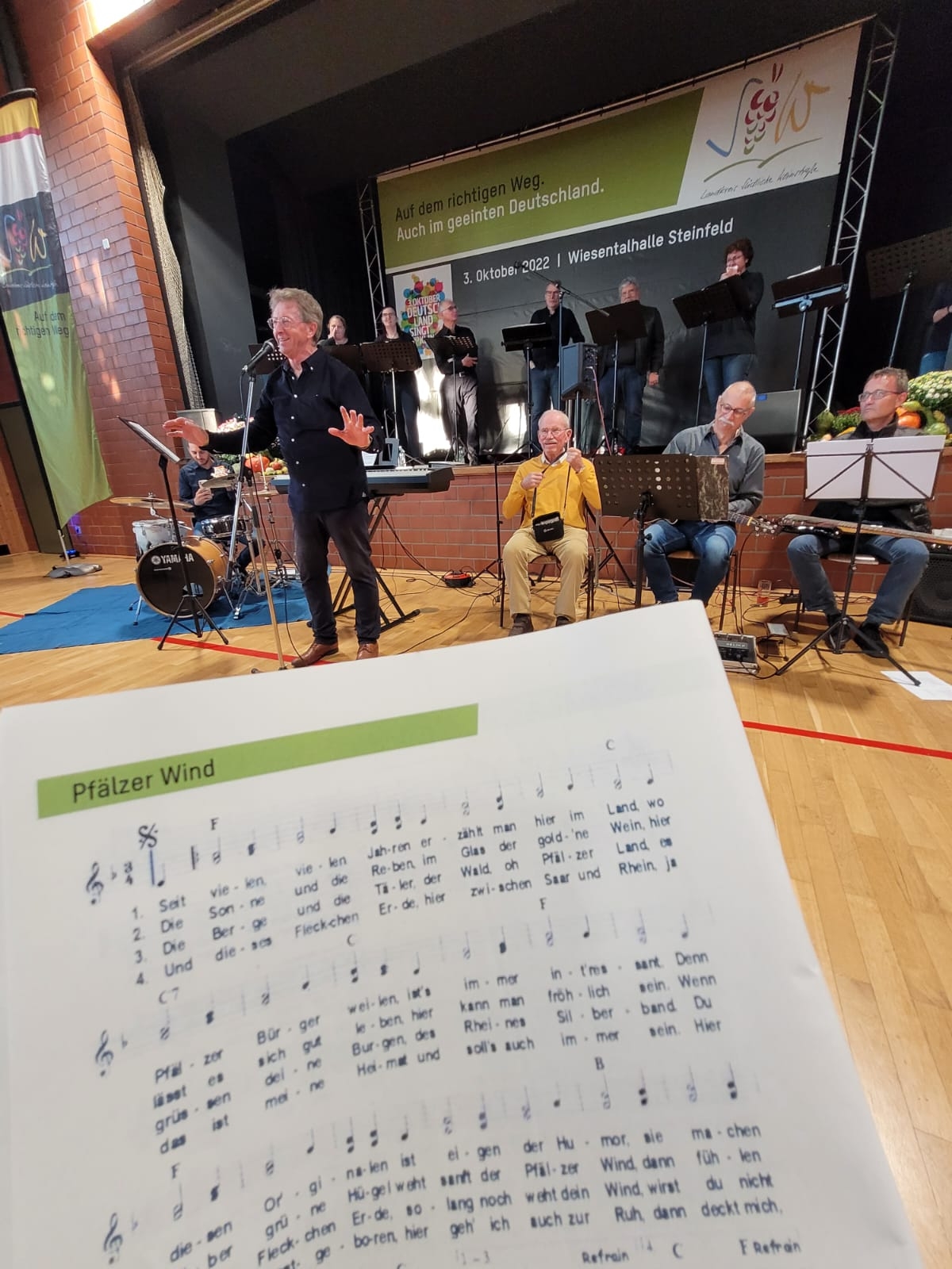 Auf dem Bild ist eine musikalische Veranstaltung im Rahmen der Aktion „Deutschland singt und klingt“ zu sehen, die am 3. Oktober 2022 in der Wiesentalhalle Steinfeld stattfand. Im Vordergrund wird ein Liederheft mit dem Titel „Pfälzer Wind“ gezeigt, während im Hintergrund eine Live-Band unter der Leitung eines Dirigenten auf der Bühne musiziert. Einige Chormitglieder singen dazu. Die Veranstaltung diente dem gemeinsamen Singen zur Feier des Tages der Deutschen Einheit. Ein Banner im Hintergrund trägt den Text „Auf dem richtigen Weg. Auch im geeinten Deutschland.
