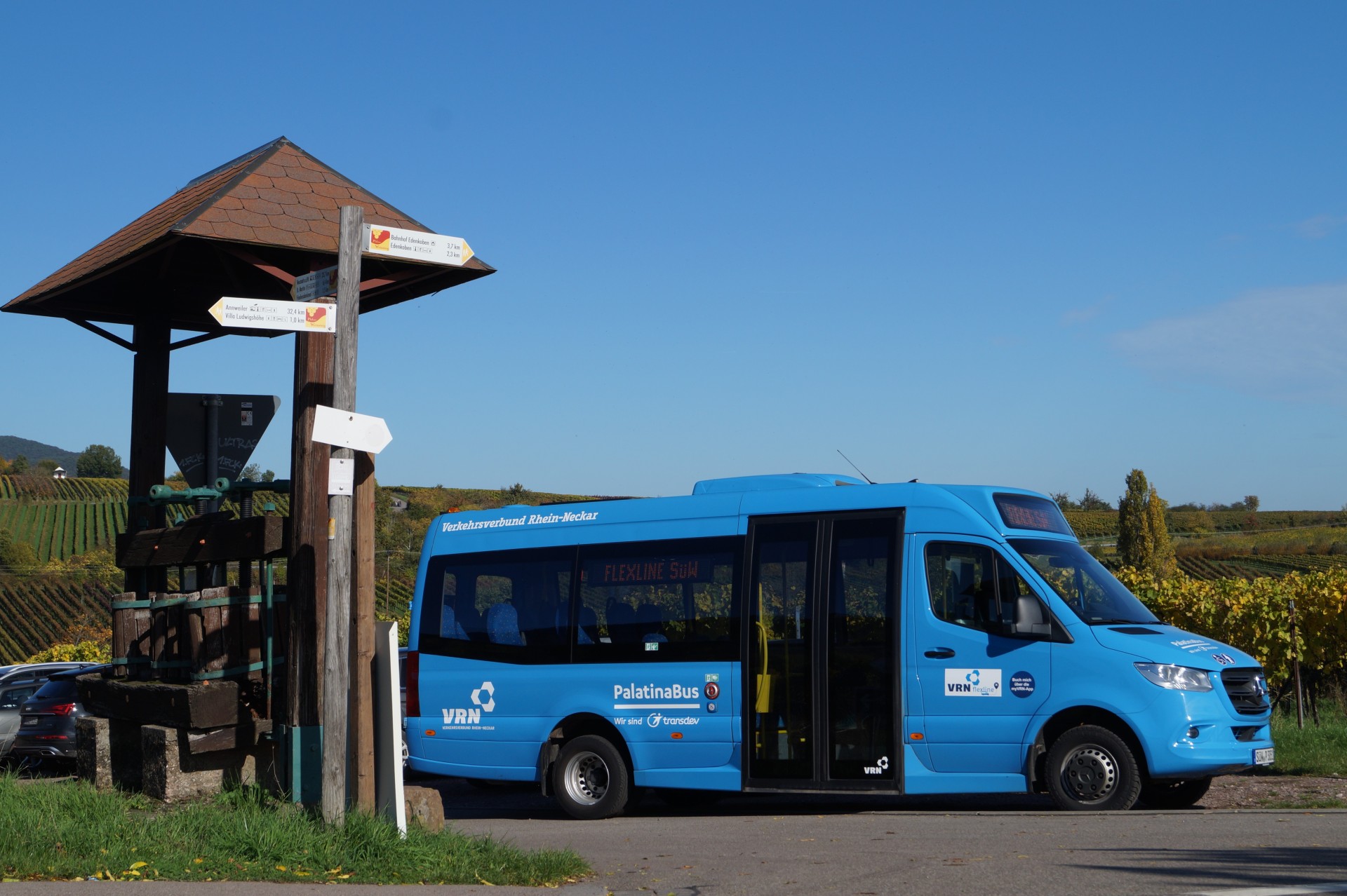 Ein blauer Kleinbus steht an einer Straßenkreuzung im ländlichen Raum. Im Hintergrund sind unter blauem Himmel Weinberge zu sehen. Auf einer digitalen Anzeigetafel im Bus ist "Flexline SÜW" zu lesen. Der Bus selbst trägt die Aufschriften "Verkehrsverbund Rhein-Neckar" und "PalatinaBus".