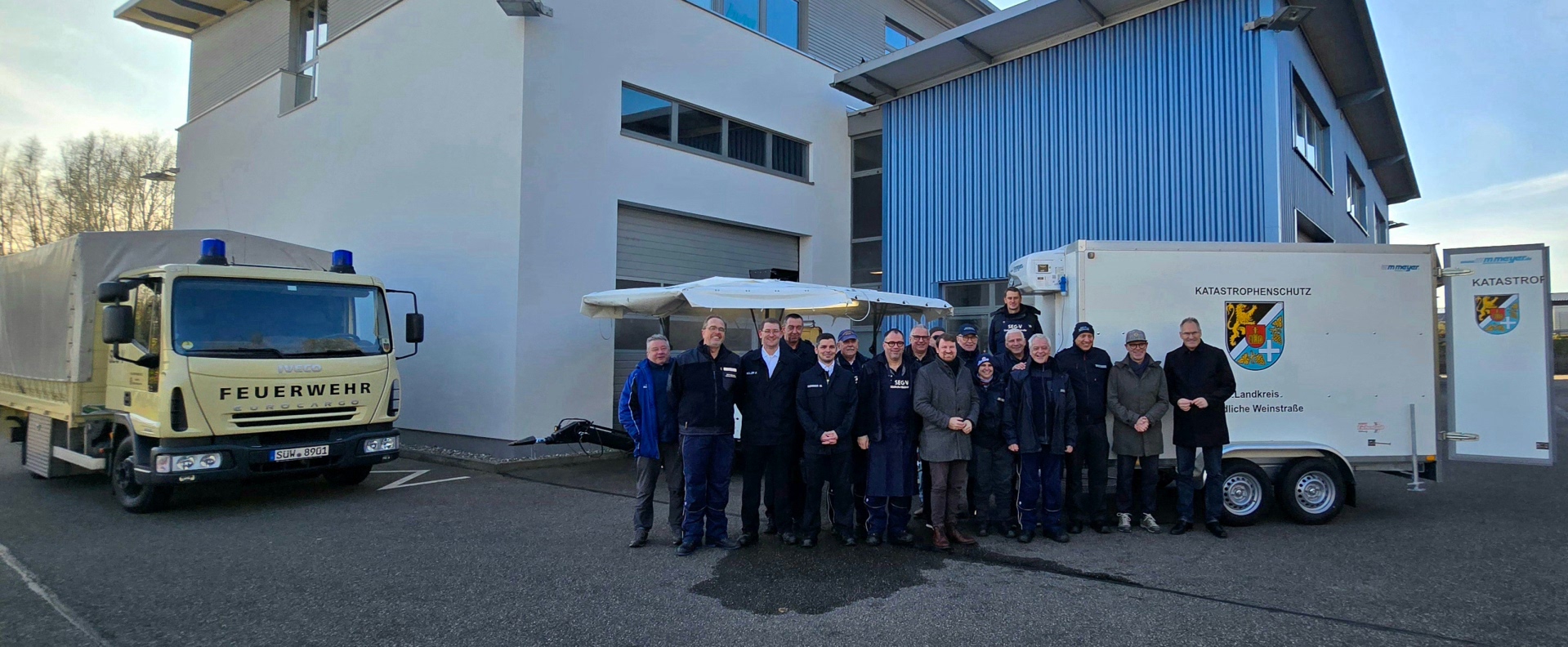 Eine Gruppe von Menschen posiert bei der Übergabe des neuen Kühlanhängers vor selbigem. Auf dem Anhänger prangt das Wappen des Landkreises SÜW unter den Schriftzug "Katastrophenschutz".