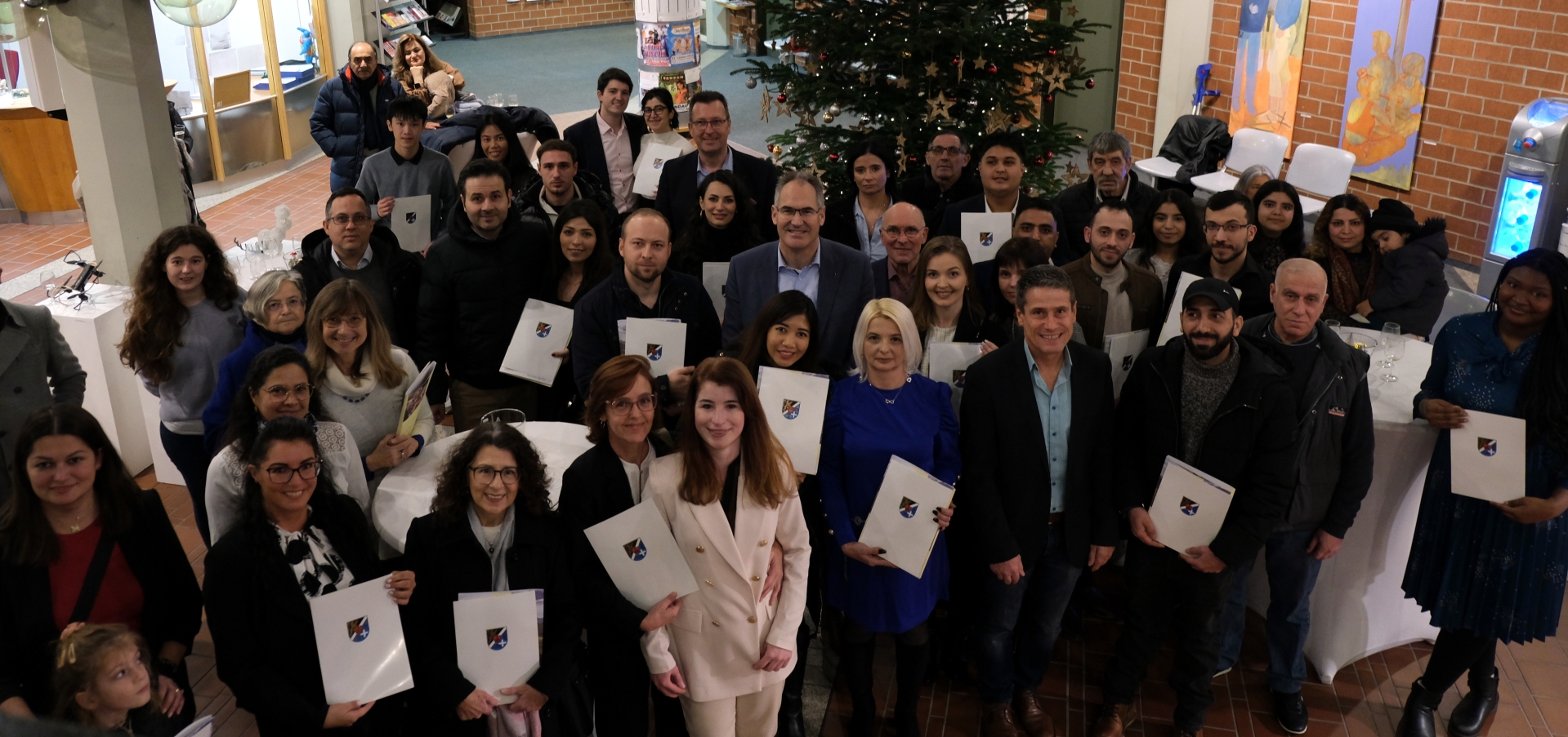 Eine Gruppe von Menschen steht bei der Einbürgerungsfeier im Foyer des Kreishauses zusammen. Im Vordergrund halten viele der Teilnehmenden ihre Einbürgerungsurkunden in der Hand. Im Hintergrund ist ein geschmückter Weihnachtsbaum sowie der Eingangsbereich des Kreishauses zu sehen.