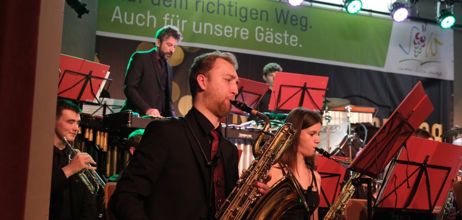 Mitglieder des Kreisjugendorchesters spielen ein Konzert. Im Vordergrund sind Musikerinnen und Musiker mit Saxophonen zu sehen, während im Hintergrund weitere Instrumente gespielt werden. Über der Bühne hängt ein Banner mit der Aufschrift „Auf dem richtigen Weg. Auch für unsere Gäste.