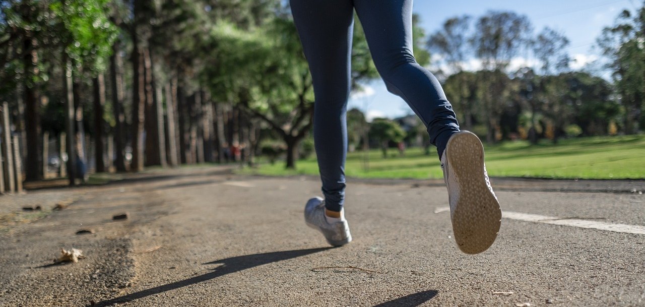 Nahaufnahme einer Person, die auf einem asphaltierten Weg in einem Park joggt. Im Hintergrund sind Bäume und eine grüne Wiese unter blauem Himmel zu sehen.
