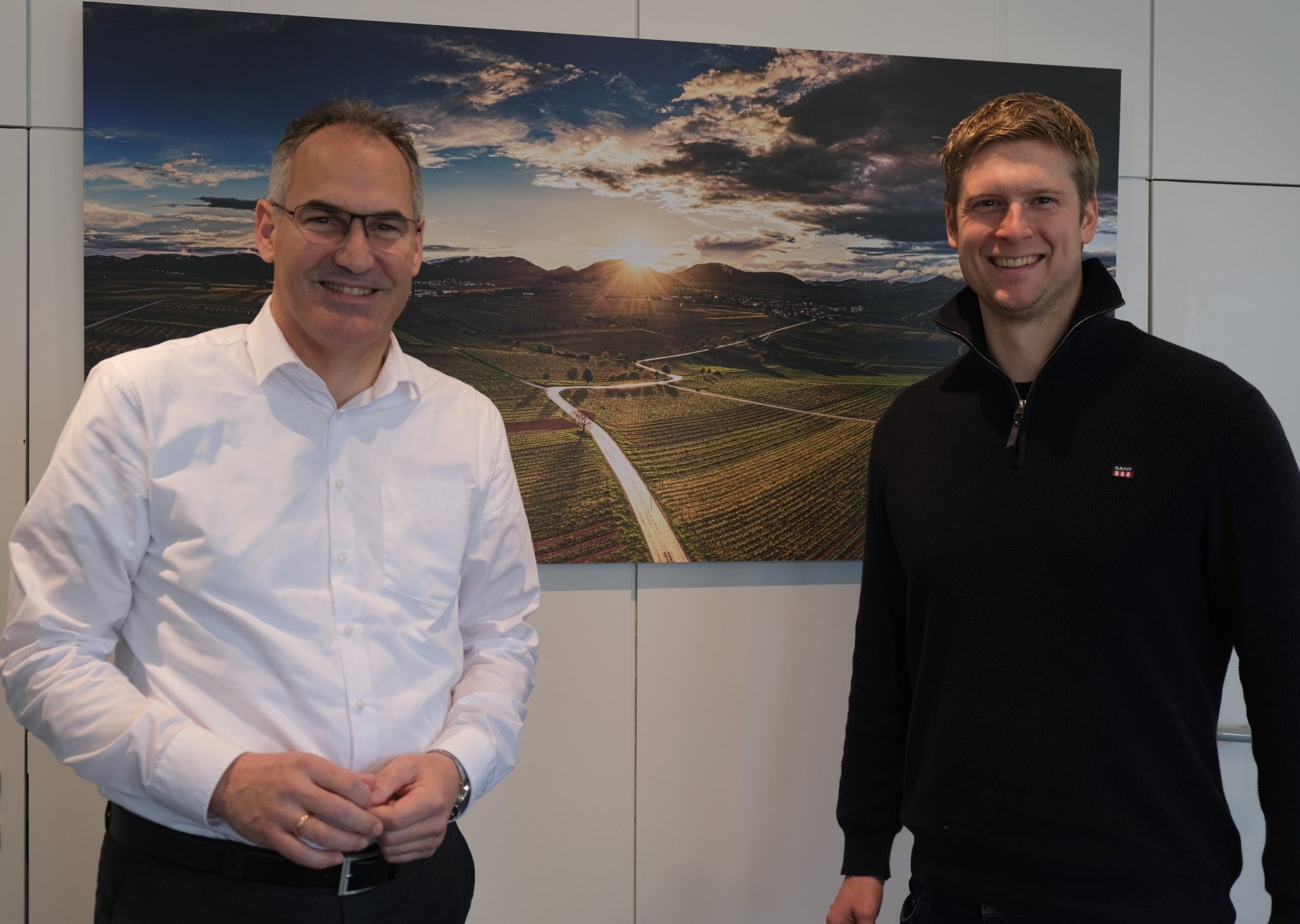 VDP.Pfalz-Vorsitzender Franz Wehrheim und Landrat Dietmar Seefeldt im Portrait. Im Hintergrund ist ein Landschaftsbild zu sehen, das die von Weinbergen geprägte Landschaft an der Südlichen Weinstraße vor den Ausläufern des Pfälzerwalds zeigt, hinter dem die Sonne untergeht.