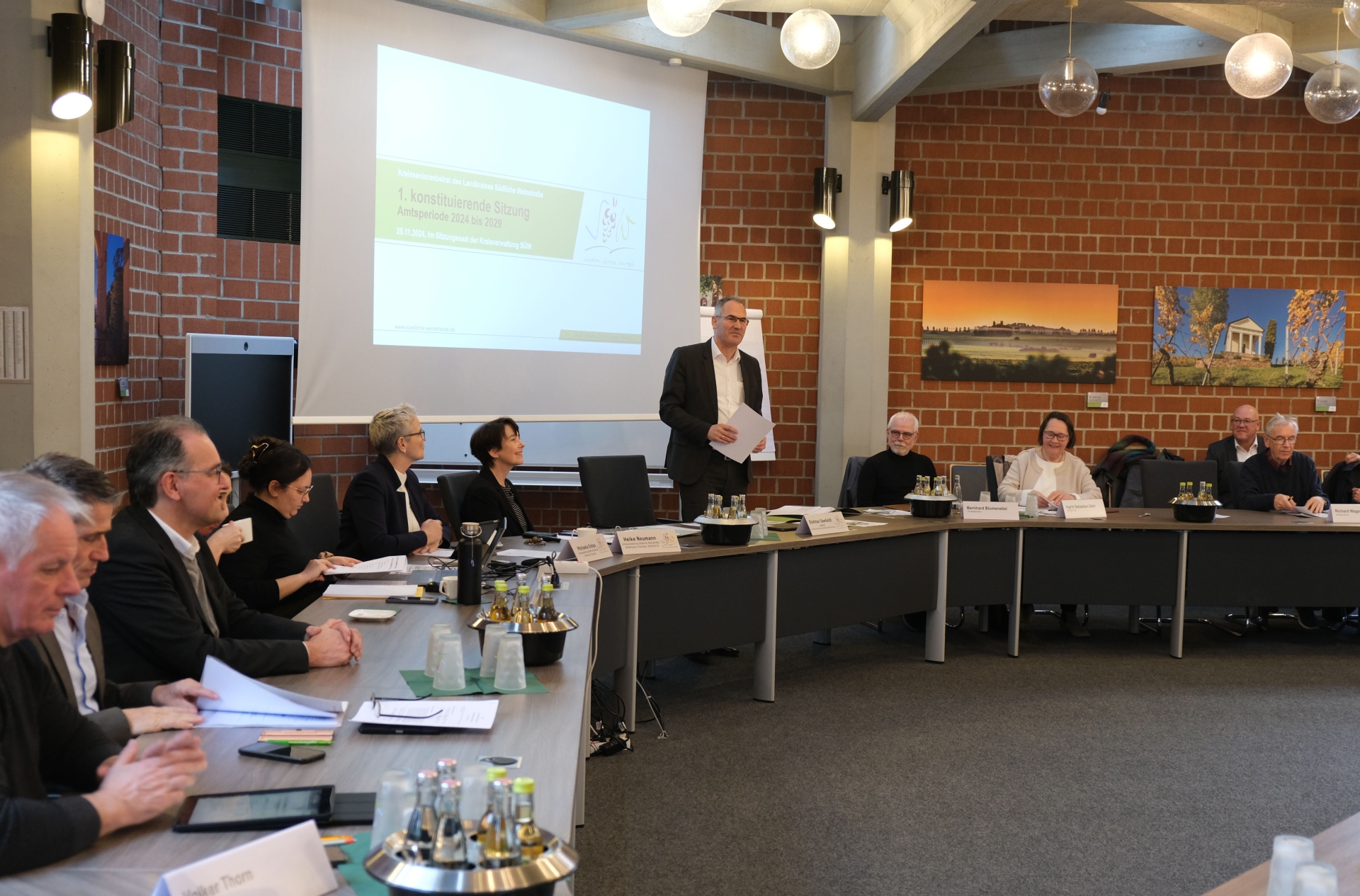 Landrat Dietmar Seefeldt steht während der konstituierenden Sitzung vor einer Präsentation im Sitzungssaal der Kreisverwaltung Südliche Weinstraße. Um ihn herum sitzen Mitglieder des Kreistags und der Verwaltung an einem großen Konferenztisch. Im Hintergrund ist eine Leinwand mit der Sitzungstagesordnung zu sehen.