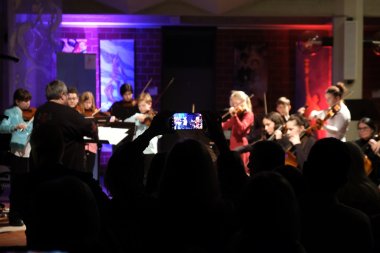 Zum Kerzenlichtkonzert der Kreismusikschule am Sonntag kamen zahlreiche Besucherinnen und Besucher in das Foyer der Kreisverwaltung SÜW. Foto: KV SÜW Ein Ensemble von jungen Musikerinnen und Musikern spielt Streichinstrumente beim Kerzenlichtkonzert der Kreismusikschule. Im Vordergrund hält eine Person ein Smartphone hoch, um das Konzert aufzunehmen. Der Hintergrund ist in stimmungsvolles farbiges Licht getaucht.