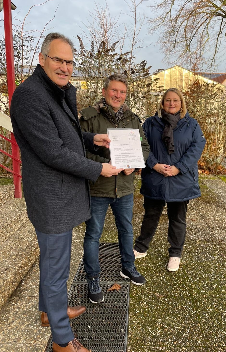 Landrat Dietmar Seefeldt (links) übergibt Ortsbürgermeister Matthias Neufeld und Beigeordneter Diana Nowak den Förderbescheid für die kommunale Kita.