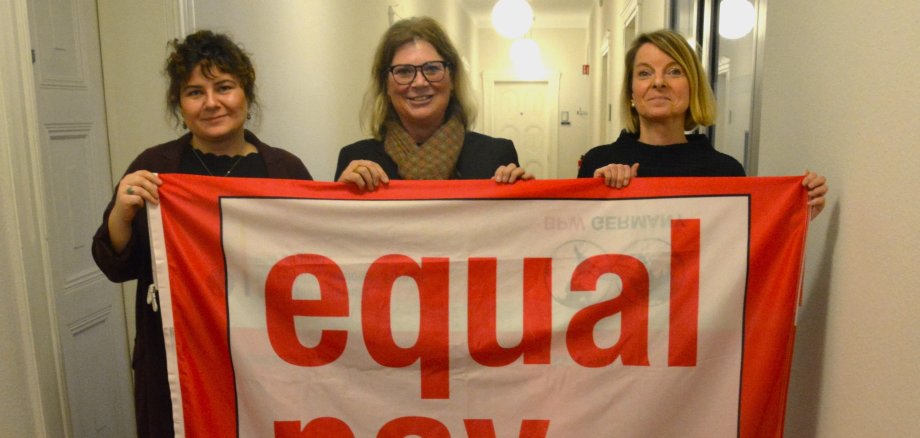 Drei Frauen halten ein rotes Banner hoch. 