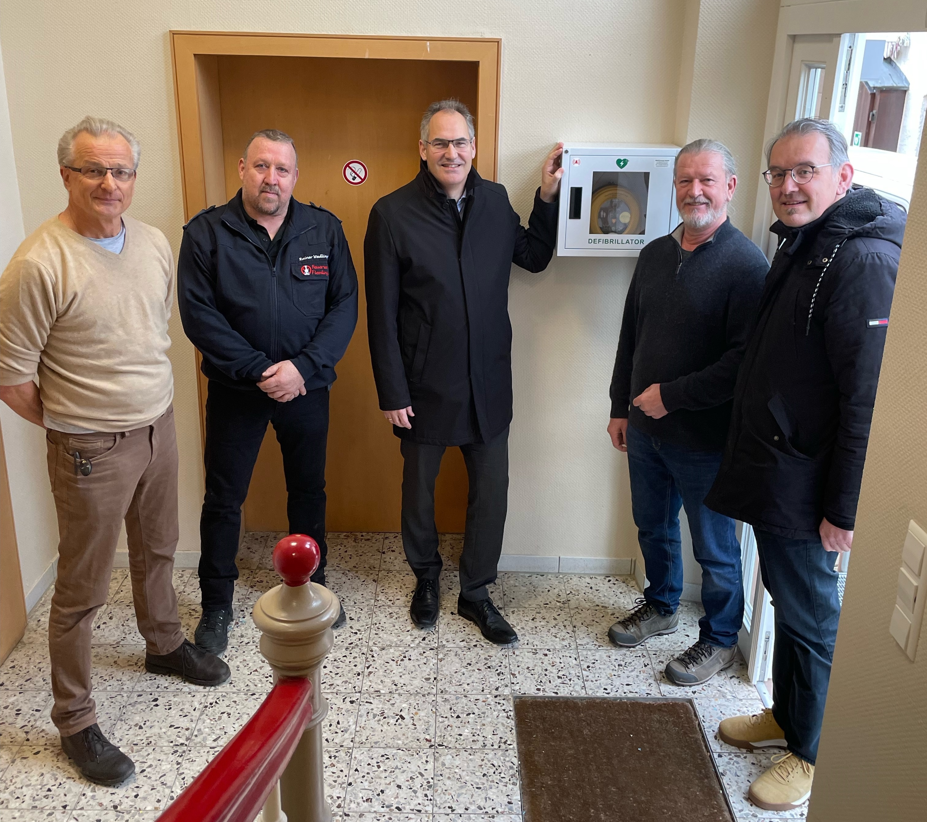 Landrat Dietmar Seefeldt zeigt bei der offiziellen Übergabe das neue AED-Gerät, das an einer Wand im Dorfgemeinschaftshaus in Flemlingen hängt. Neben ihm stehen Ortsbeigeordneter Georg Minges, Wehrführer Rainer Wadlinger, Ortsbürgermeister Peter Henrich und Erster Kreisbeigeordneter Georg Kern.