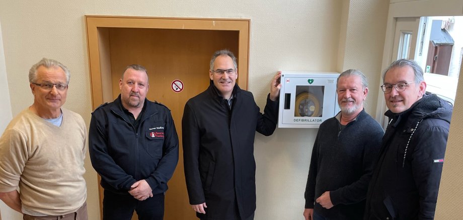 Landrat Dietmar Seefeldt zeigt bei der offiziellen Übergabe das neue AED-Gerät, das an einer Wand im Dorfgemeinschaftshaus in Flemlingen hängt. Neben ihm stehen Ortsbeigeordneter Georg Minges, Wehrführer Rainer Wadlinger, Ortsbürgermeister Peter Henrich und Erster Kreisbeigeordneter Georg Kern.