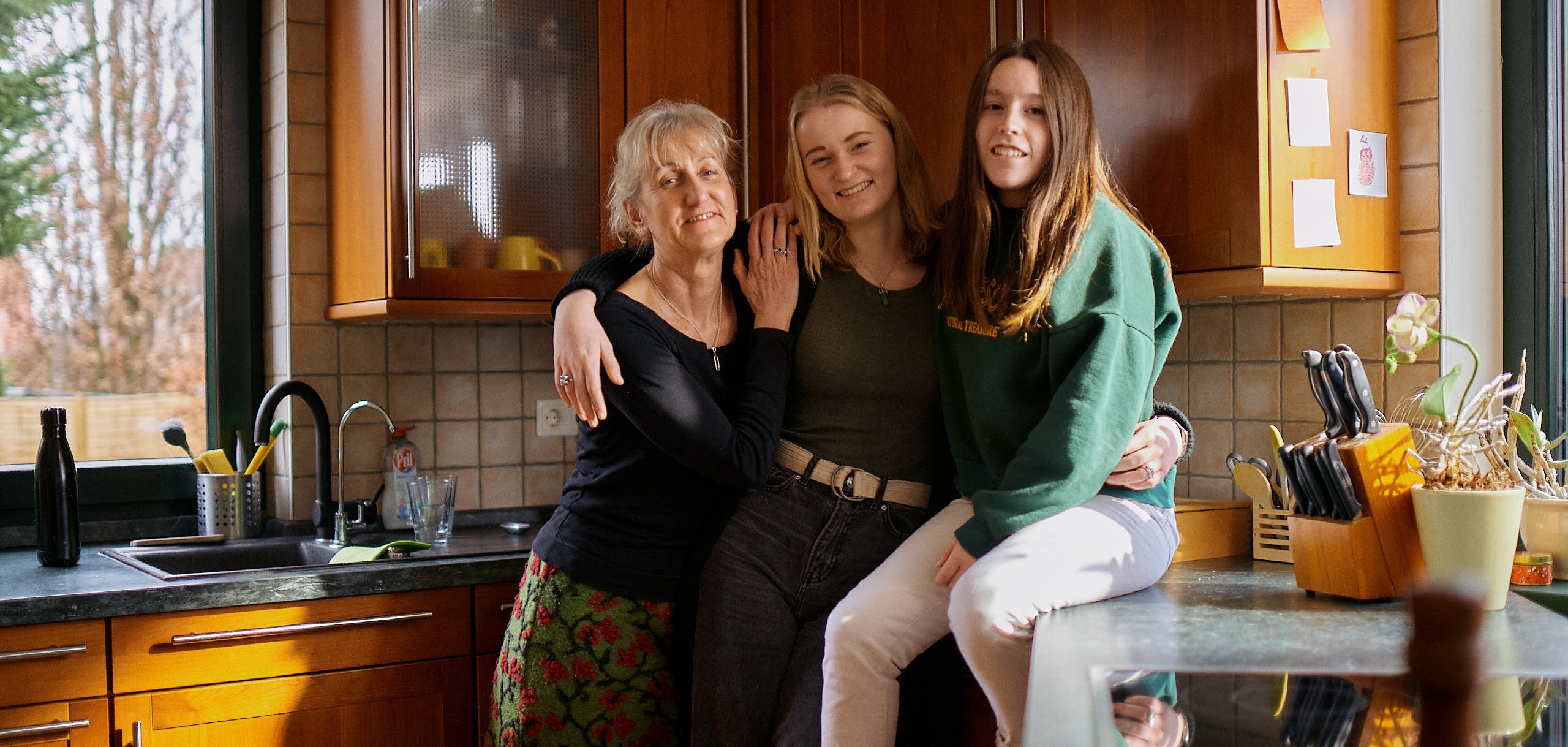 Eine erwachsene Frau und zwei Jugendliche lehnen sich aneinander, eine der Jugendlichen sitzt auf der Küchenarbeitsfläche. Foto: AFS