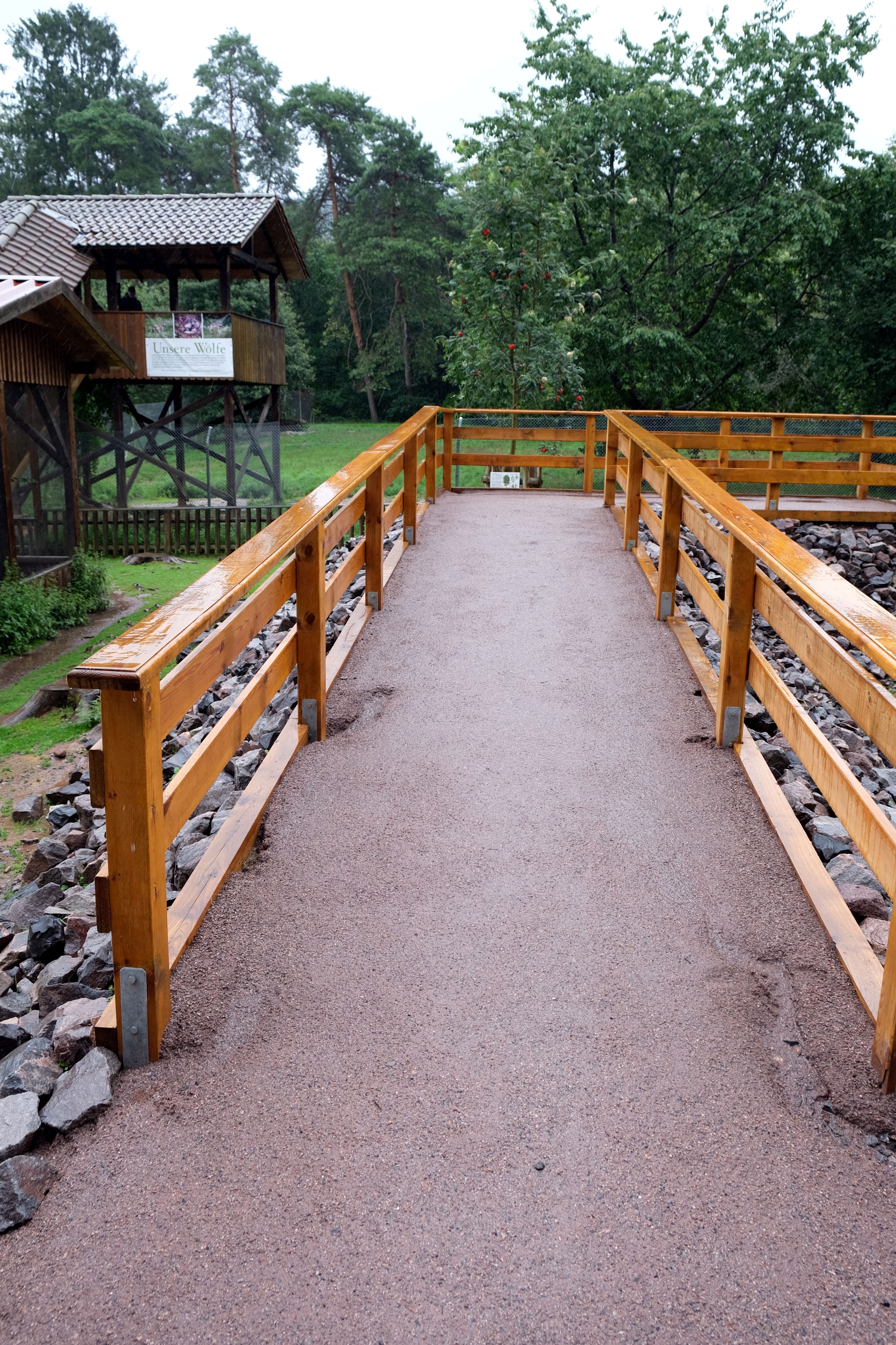 Ein barrierefreier Weg mit Holzgeländer führt zum Wolfsgehege im Wild- und Wanderpark. Im Hintergrund sind Bäume und ein Gebäude mit einem Schild 'Unsere Wölfe' zu sehen.