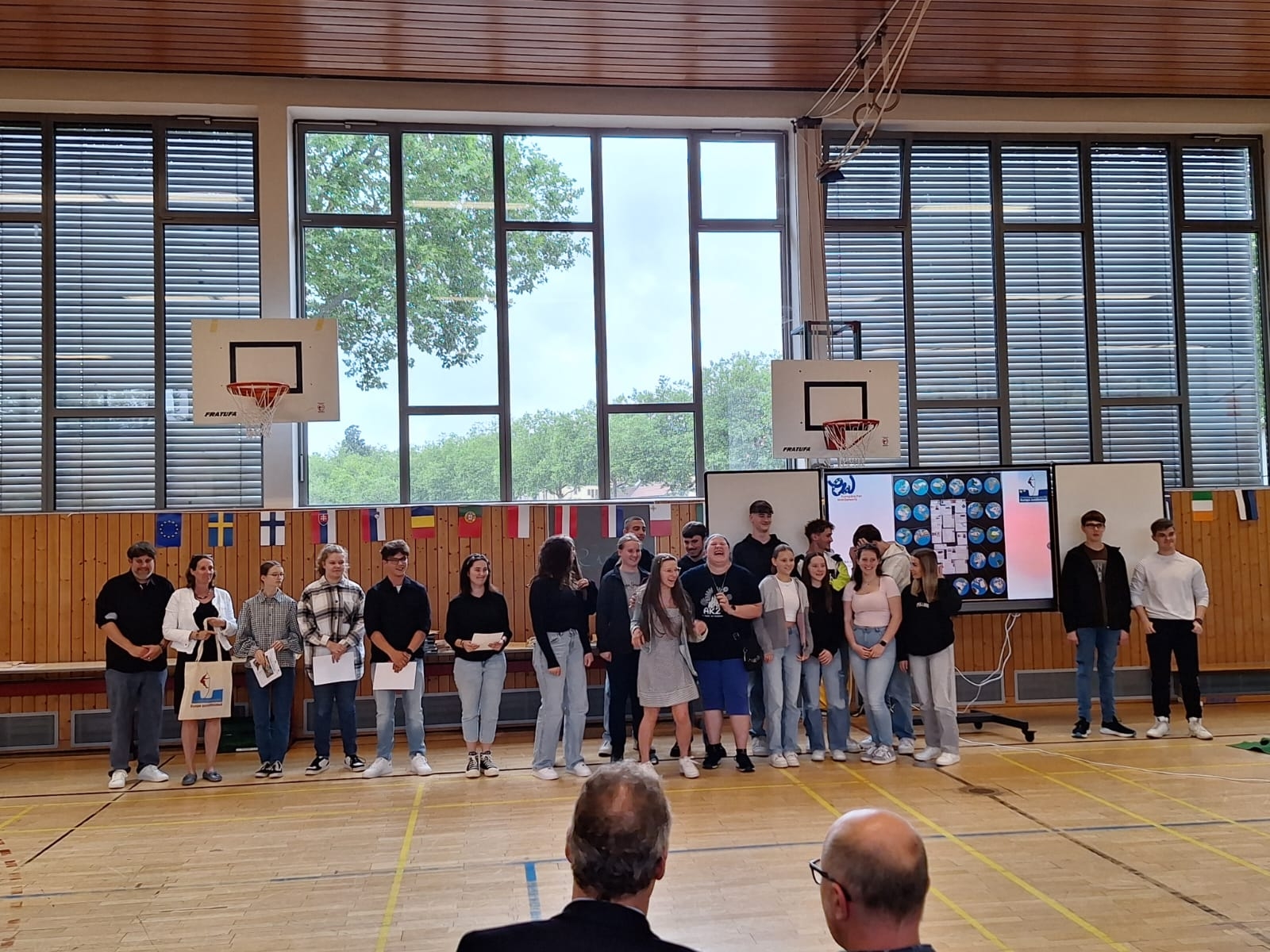 Schülerinnen und Schüler stehen in einer Turnhalle, dahinter verschiedene Nationalflaggen.