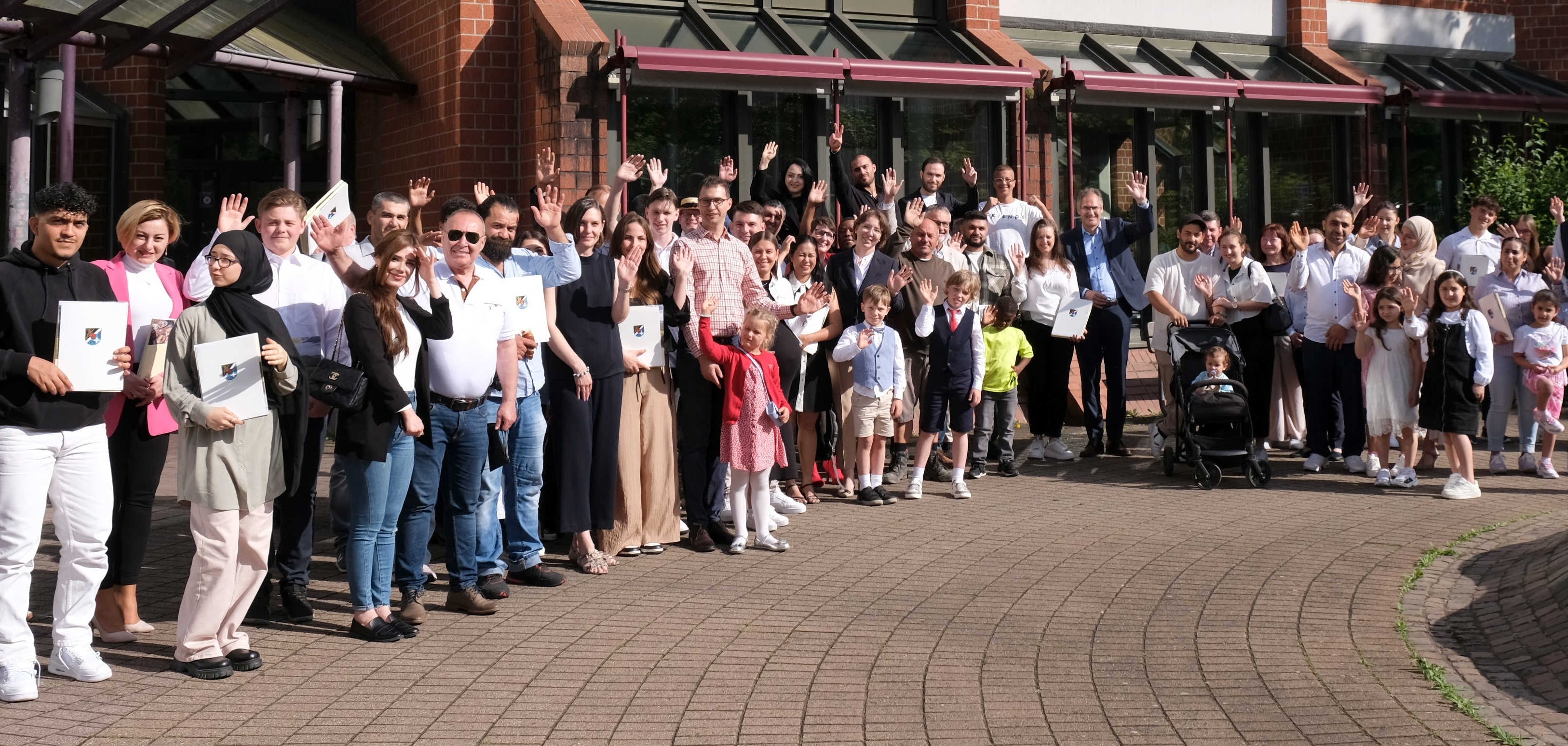 Eine große Gruppe von Menschen, darunter Erwachsene und Kinder, steht vor dem Kreishaus und lächelt in die Kamera. Viele halten Dokumentenmappen in der Hand. Die Szene ist bei der jüngsten Einbürgerungsfeier aufgenommen.