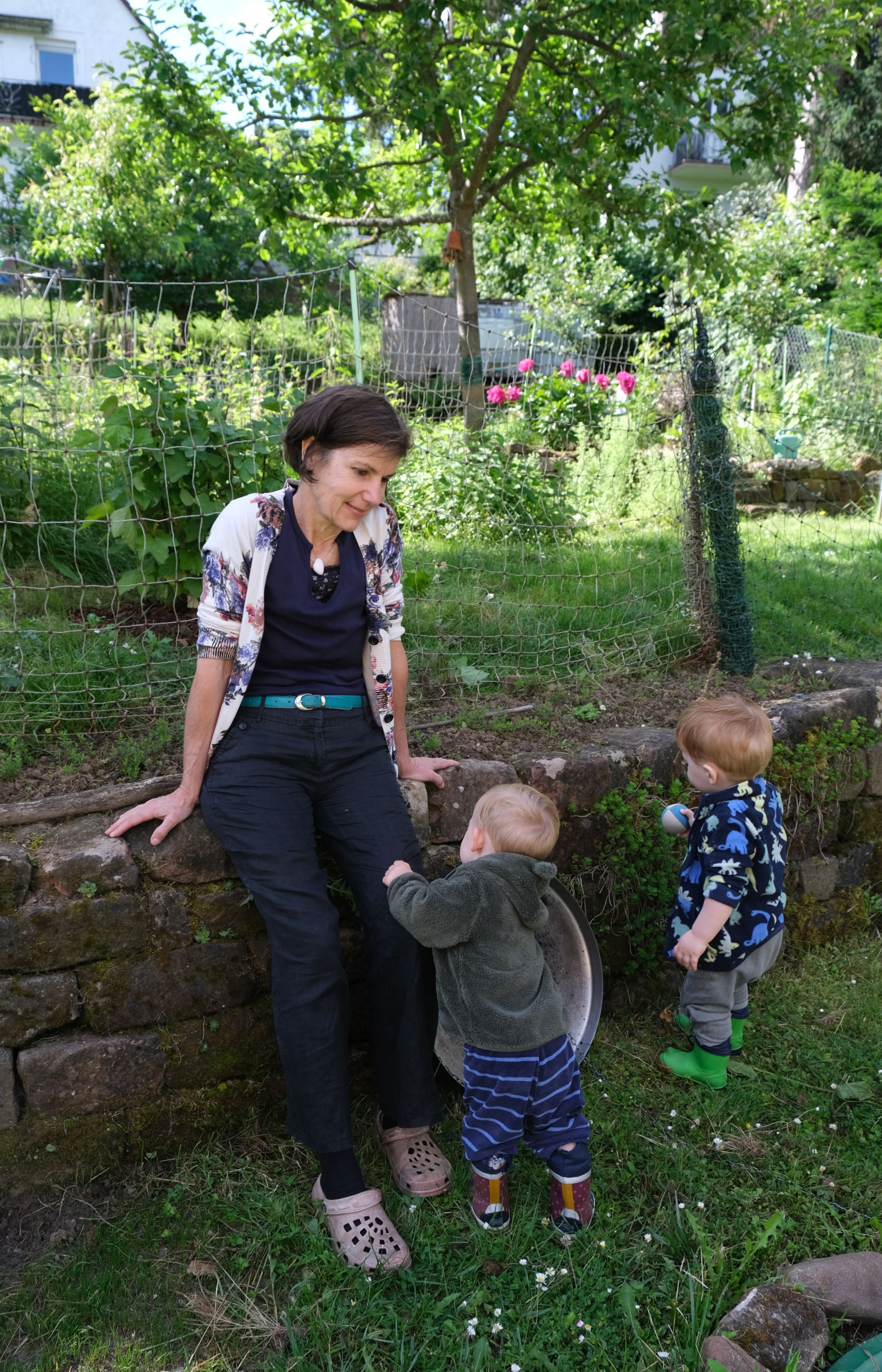 Tagesmutter Friederike Carius sitzt im Garten auf einer Mauer und beaufsichtigt zwei kleine Kinder.