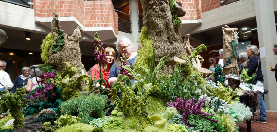 Diese Aufnahme zeigt eine Gruppe von Menschen, die eine kunstvolle, gehäkelte Waldlandschaft in einer Ausstellung im Kreishaus Südliche Weinstraße (SÜW) betrachten. Die Installation, die üppige grüne und violette Pflanzen und Bäume aus verschiedenen Wollsorten und Häkeltechniken darstellt, zieht die Aufmerksamkeit der Besucherinnen und Besucher auf sich. Im Hintergrund sind weitere Ausstellungsbesucher zu sehen.
