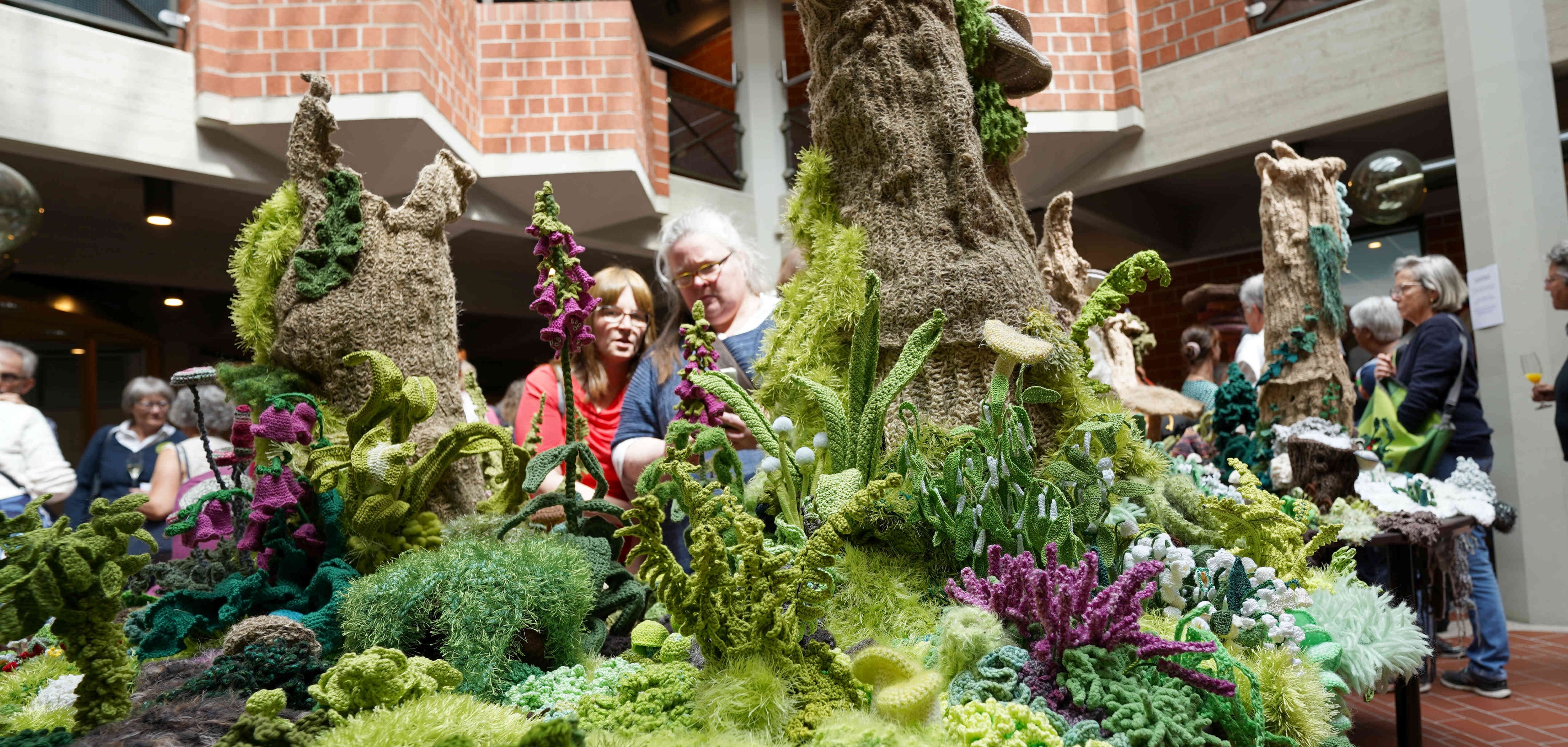 Diese Aufnahme zeigt eine Gruppe von Menschen, die eine kunstvolle, gehäkelte Waldlandschaft in einer Ausstellung im Kreishaus Südliche Weinstraße (SÜW) betrachten. Die Installation, die üppige grüne und violette Pflanzen und Bäume aus verschiedenen Wollsorten und Häkeltechniken darstellt, zieht die Aufmerksamkeit der Besucherinnen und Besucher auf sich. Im Hintergrund sind weitere Ausstellungsbesucher zu sehen.