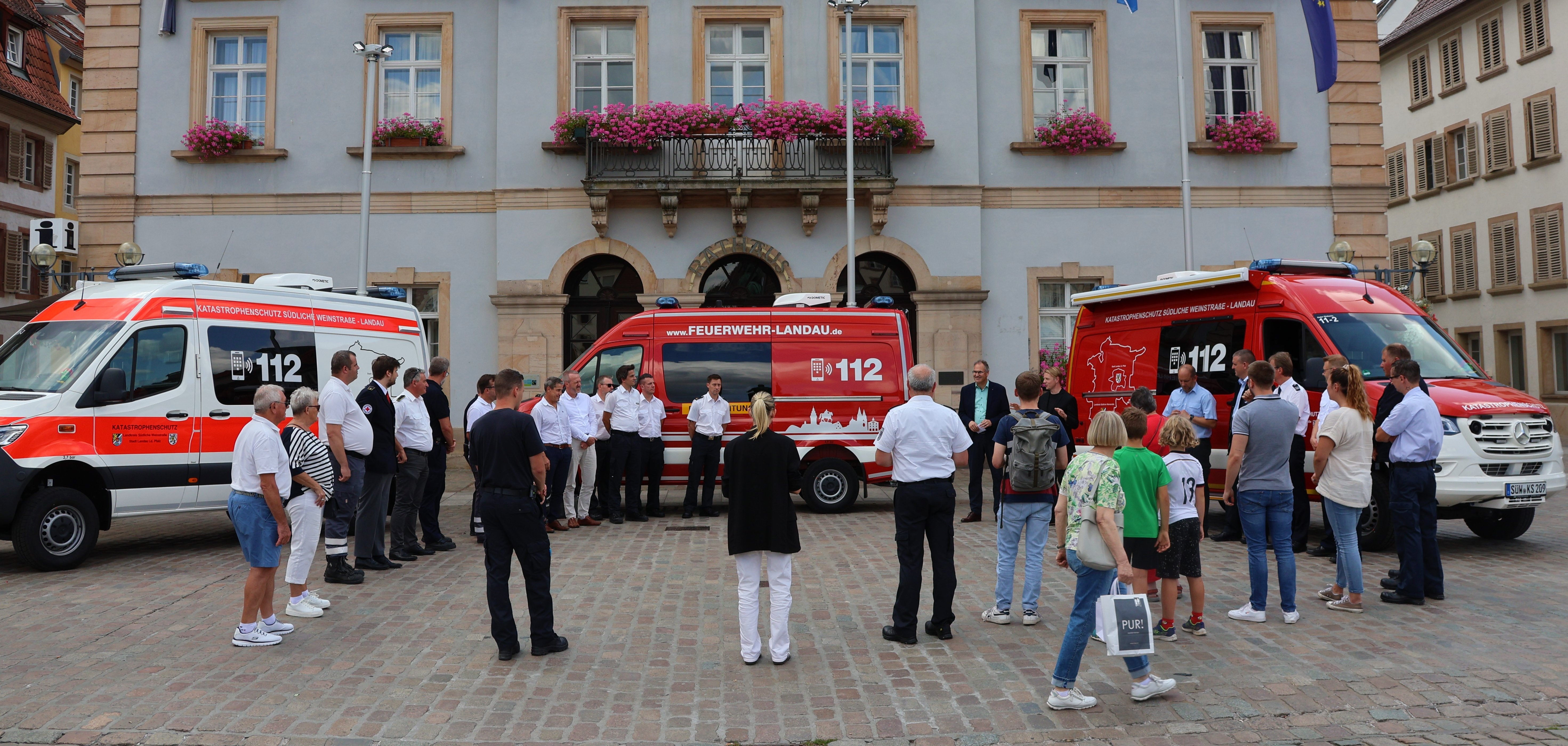 Eine Gruppe von Feuerwehrleuten und Mitgliedern des Katastrophenschutzes steht vor dem historischen Gebäude des Rathauses in Landau. Vor ihnen sind drei neue Einsatzleitwagen der Feuerwehr Landau und des Katastrophenschutzes des Landkreises Südliche Weinstraße geparkt. Die Einsatzfahrzeuge sind modern und in auffälligen Farben gestaltet. Blumen schmücken die Balkone des Rathauses, während Fahnen im Wind wehen.
