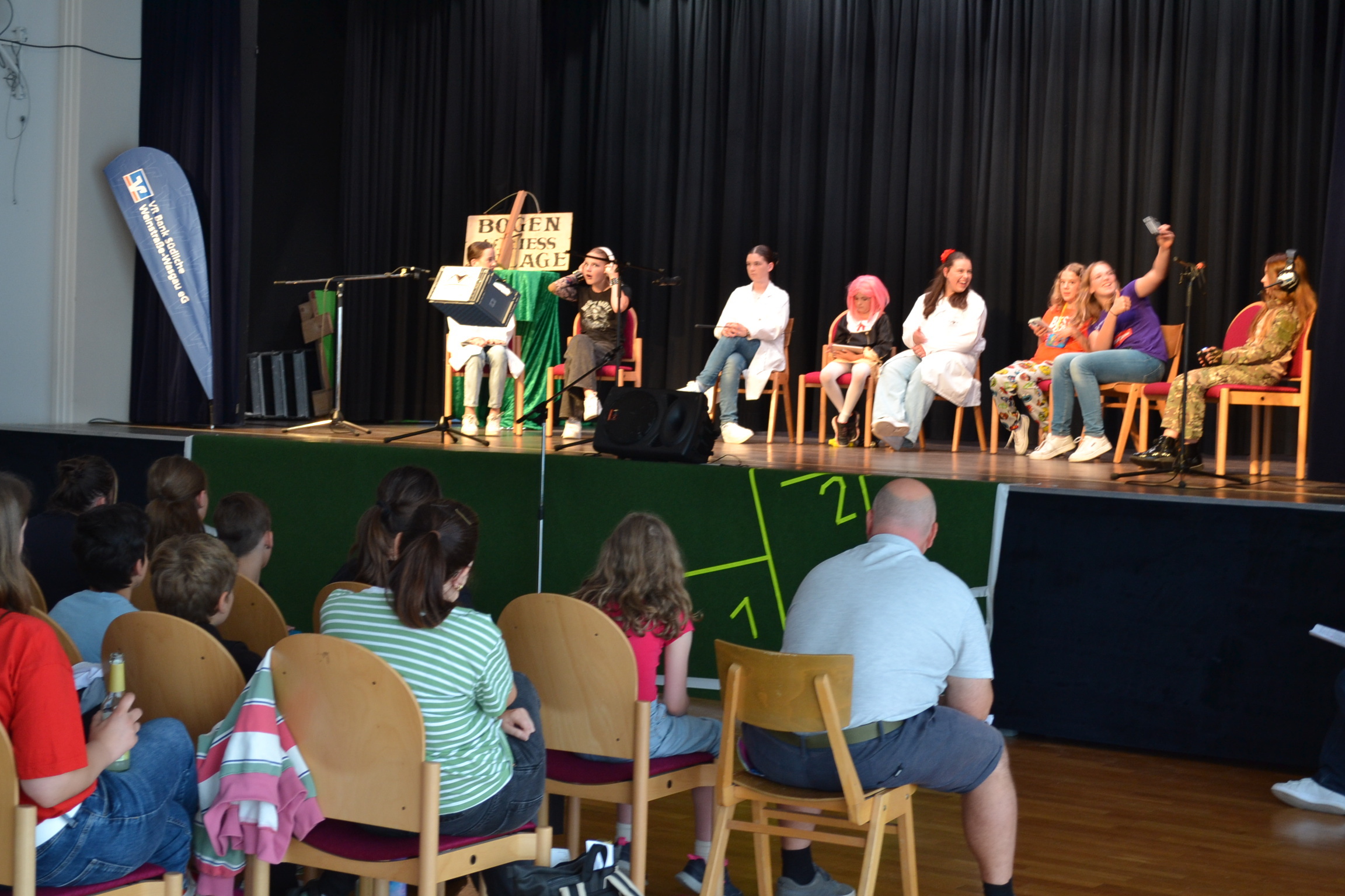 Eine Bühne mit acht jungen Menschen in verschiedenen Kostümen, die auf Stühlen sitzen und an einer Theatervorführung oder einem Bühnenprogramm teilnehmen. Im Vordergrund des Bildes sitzt ein Publikum, das aus Kindern und Erwachsenen besteht.