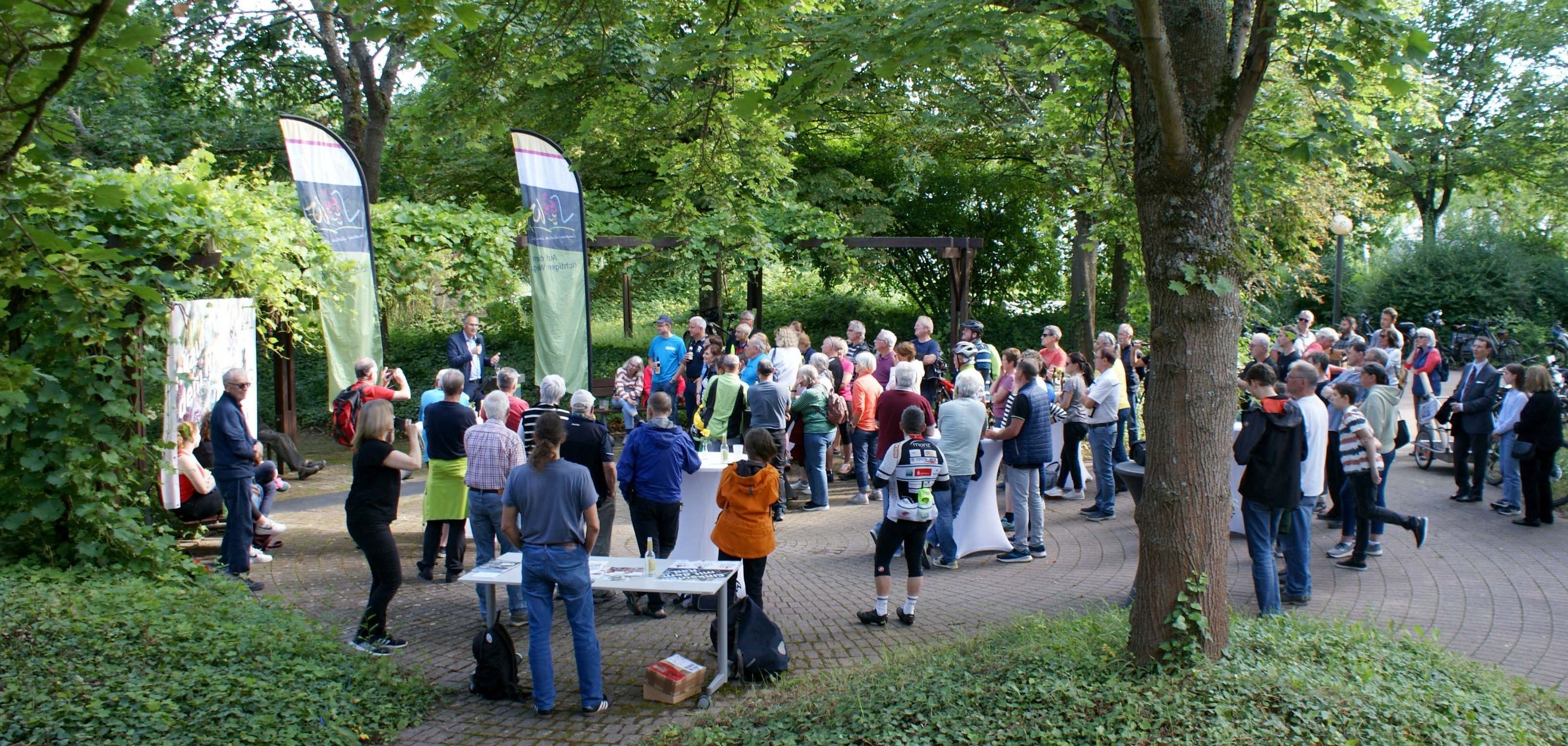 Eine Versammlung von über 120 Personen im Freien unter einem Baumdach. Die Menschen stehen am Ziel der Sternfahrt im Rahmen des STADTRADELN-Events, organisiert von der Kreisverwaltung Südliche Weinstraße (SÜW). Die Teilnehmer stehen oder sitzen in Gruppen zusammen und hören einer Person zu, die vorne spricht.