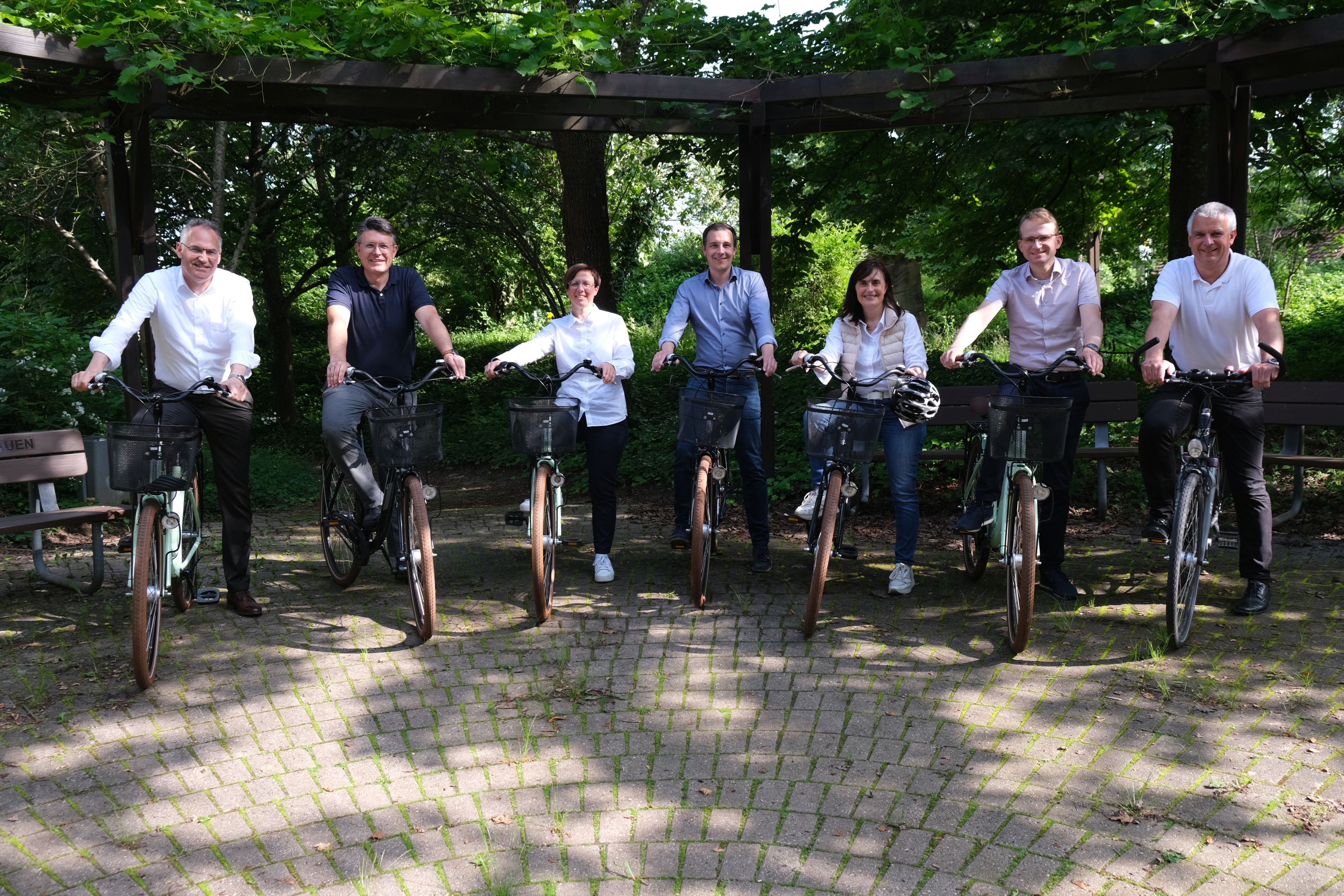 Landrat Dietmar Seefeldt sowie die Bürgermeisterinnen und Bürgermeister der sieben Verbandsgemeinden im Landkreis (Ausnahme: Offenbach an der Queich) stehen nebeneinander, jeder hat ein Fahrrad.