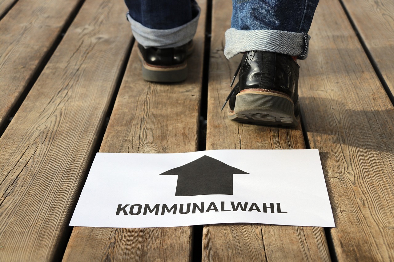 Abgebildet sind zwei Schuhe von einer laufenden Person auf Holzdielen. Im Vordergrund ein Blatt Papier mit einem Pfeil und dem Wort "Kommunalwahl" darauf.