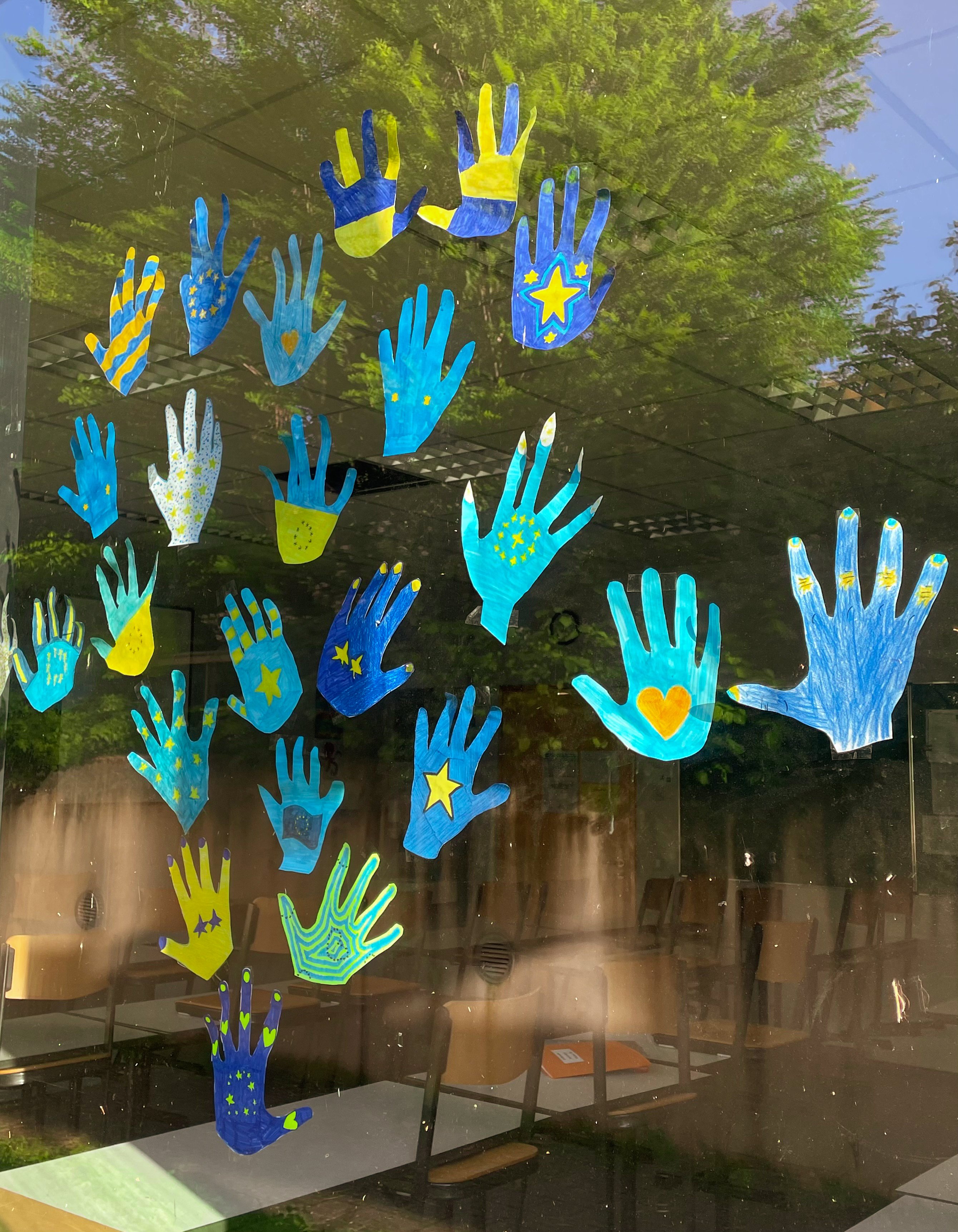 Mehrere ausgeschnittene Handabdrücke in Blau- und Gelbtönen sind an einem Fenster angebracht. Einige der Handabdrücke sind mit Sternen und Herzen verziert, andere haben Streifen oder Punkte. Die Gestaltung der Handabdrücke zeigt kreative und farbenfrohe Muster. Durch das Fenster ist der Innenraum eines Klassenzimmers mit Tischen und Stühlen zu sehen.