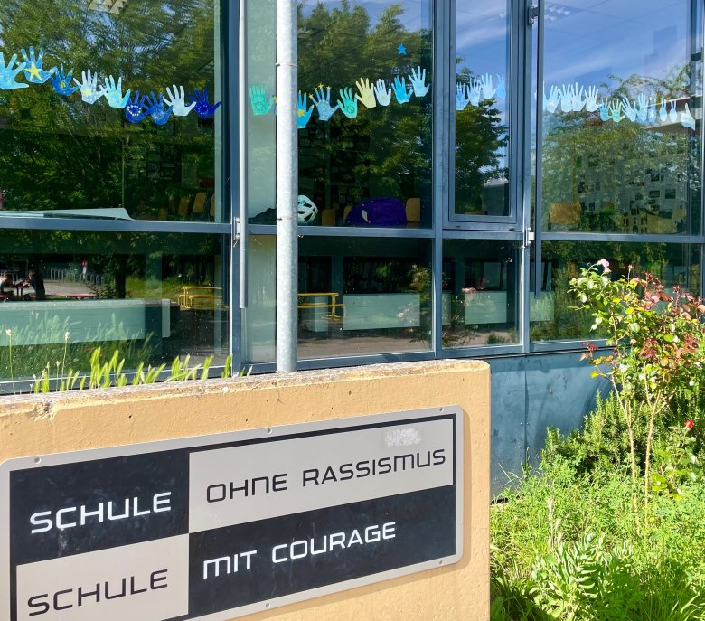 Zahlreiche Schülerinnen und Schüler haben kurz vor der Europawahl ein Zeichen für Europa sowie für die Demokratie gesetzt und die Fenster mit ihren in den EU-Farben blau und gelb kolorierten Handabdrücken geschmückt. Foto: Gymnasium Edenkoben Ein Schulschild aus Metall zeigt die Aufschrift 'Schule ohne Rassismus, Schule mit Courage'. Das Schild ist an einer Wand angebracht, vor der sich eine Glasfassade befindet. Hinter der Glasfassade sind bunte, ausgeschnittene Handabdrücke zu sehen, die an der Innenseite der Fenster angebracht sind. Die Handabdrücke sind in Blau- und Gelbtönen gestaltet. Im Hintergrund ist ein Klassenzimmer mit leeren Stühlen zu erkennen.